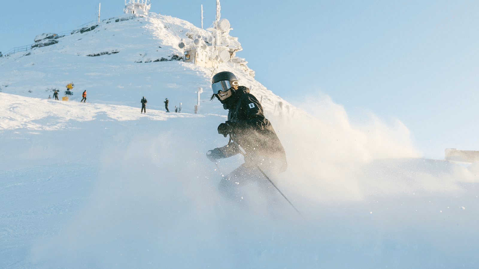 Skier in black gear and helmet skiing down a snowy mountain with snow spray around.