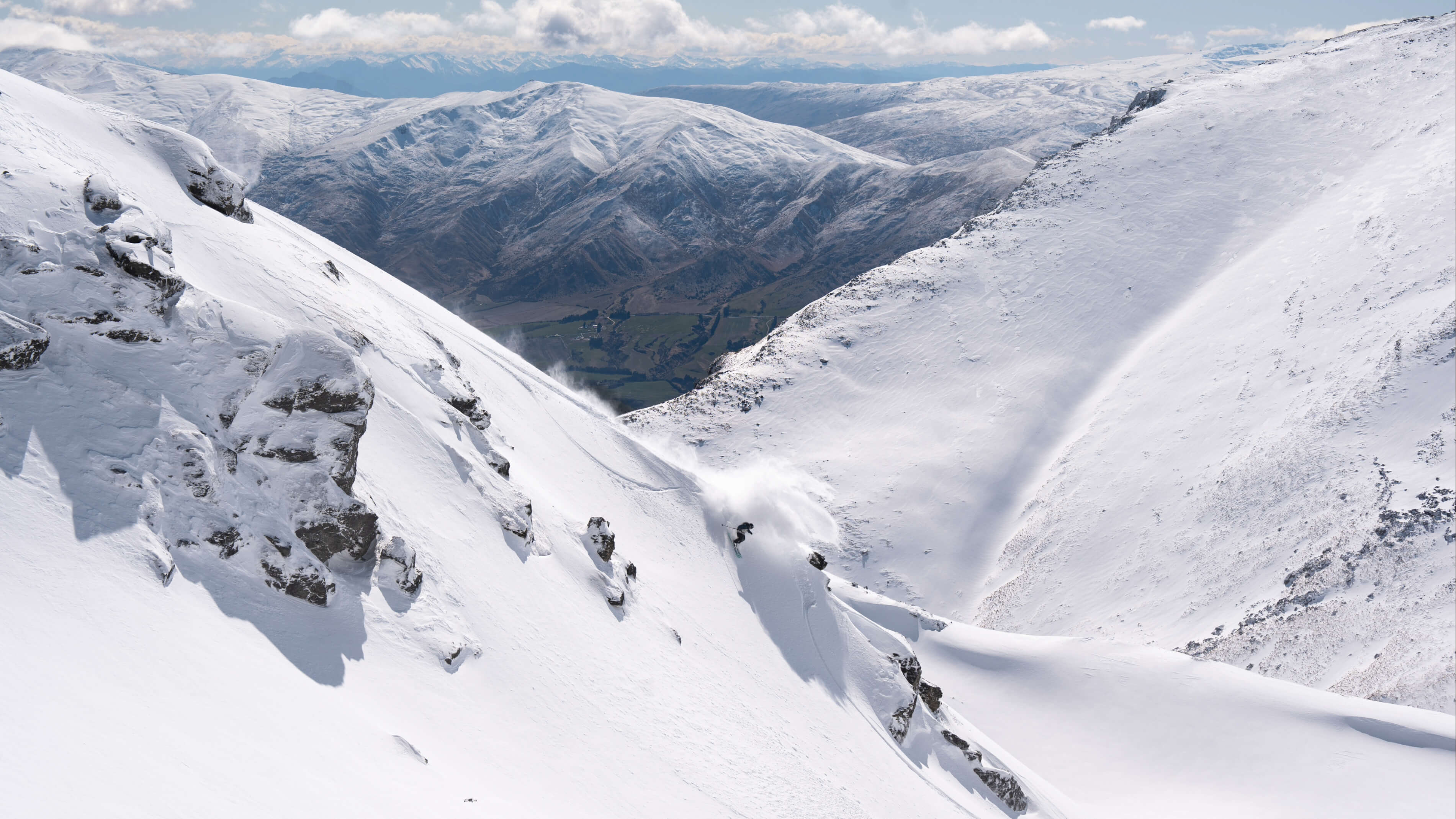 Freeride skier making a turn down a snow-covered mountain slope with snowy mountains in the background.
