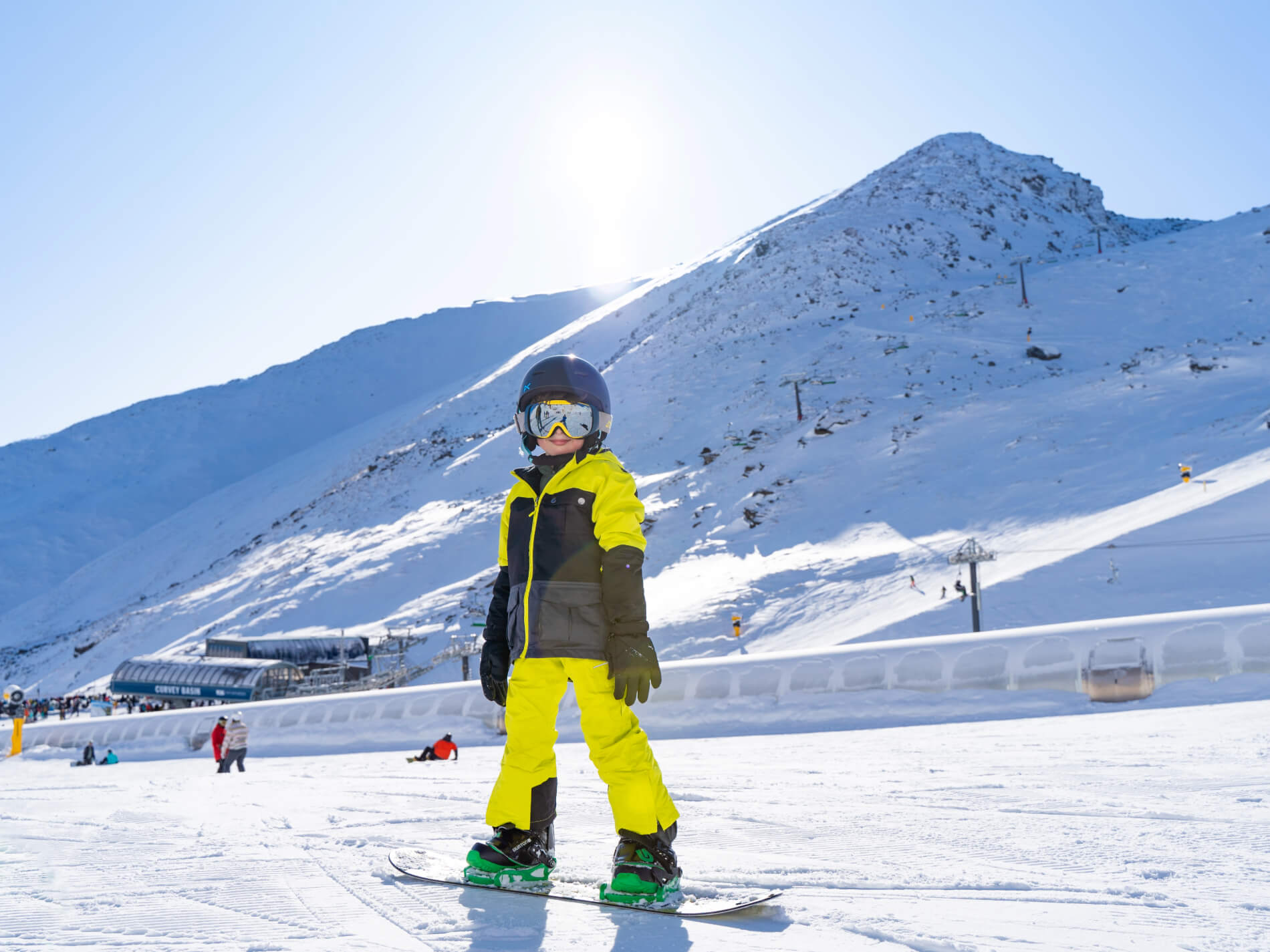 Child in yellow ski outfit and helmet snowboarding on a beginner slope with snowy mountains behind.