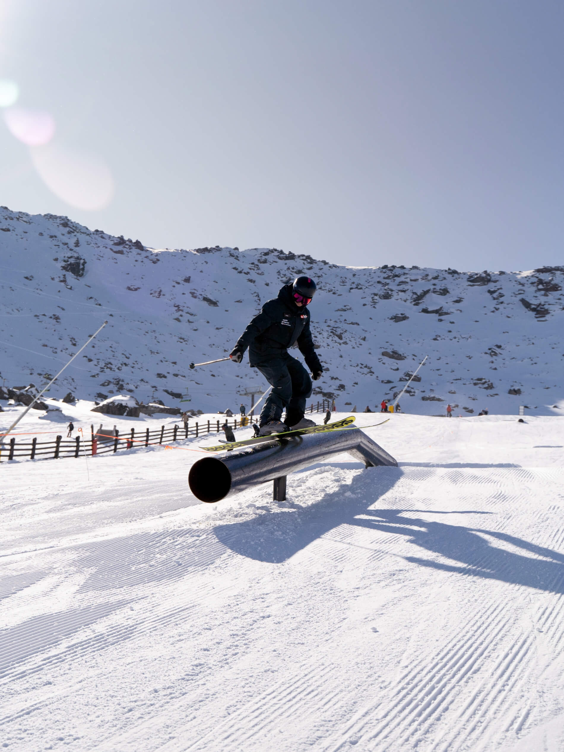 Skier in black gear sliding on a metal rail in a snowy ski park with mountains in the background.