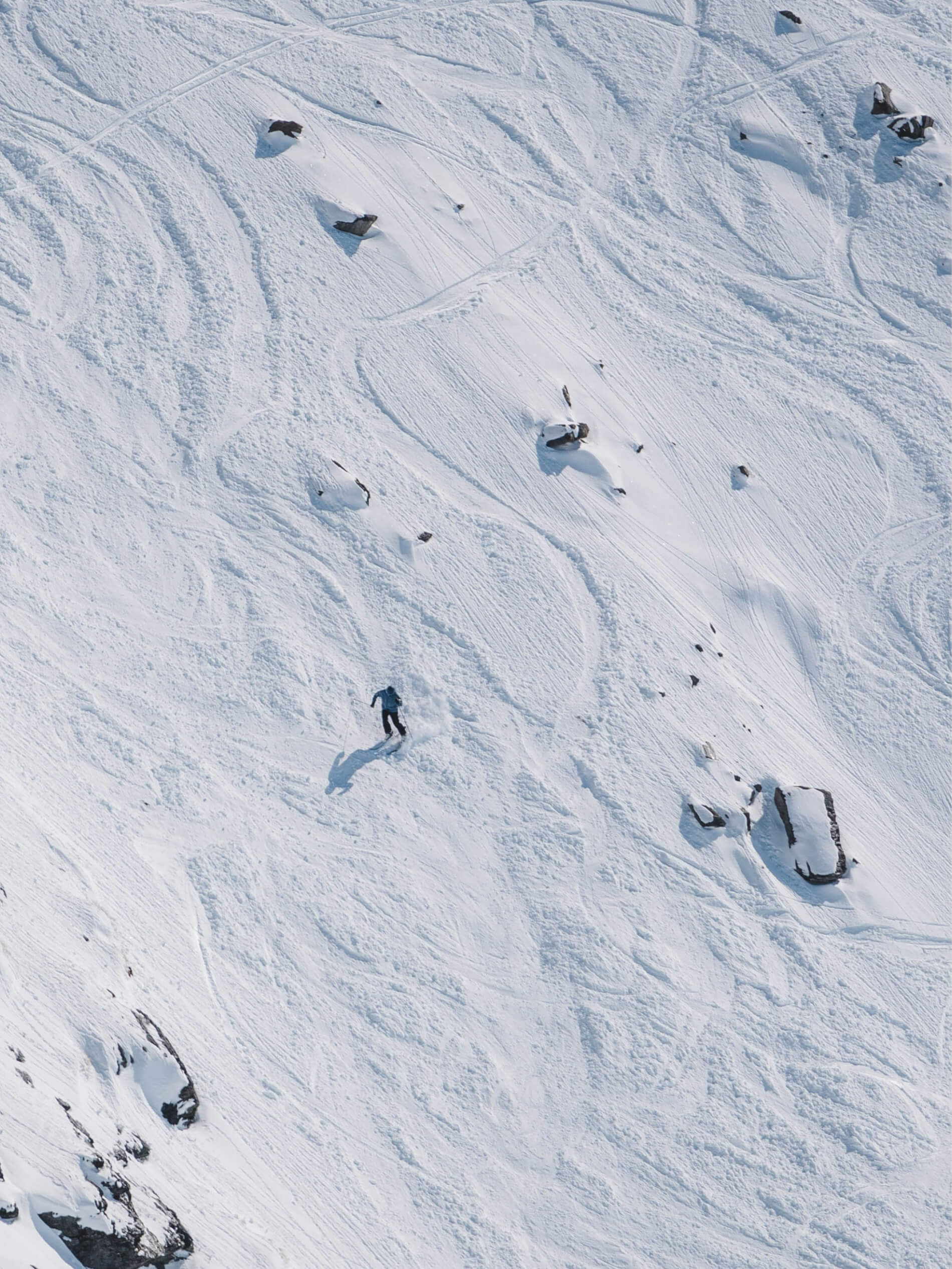A lone skier descending a snowy mountain slope with scattered rocks and ski tracks.