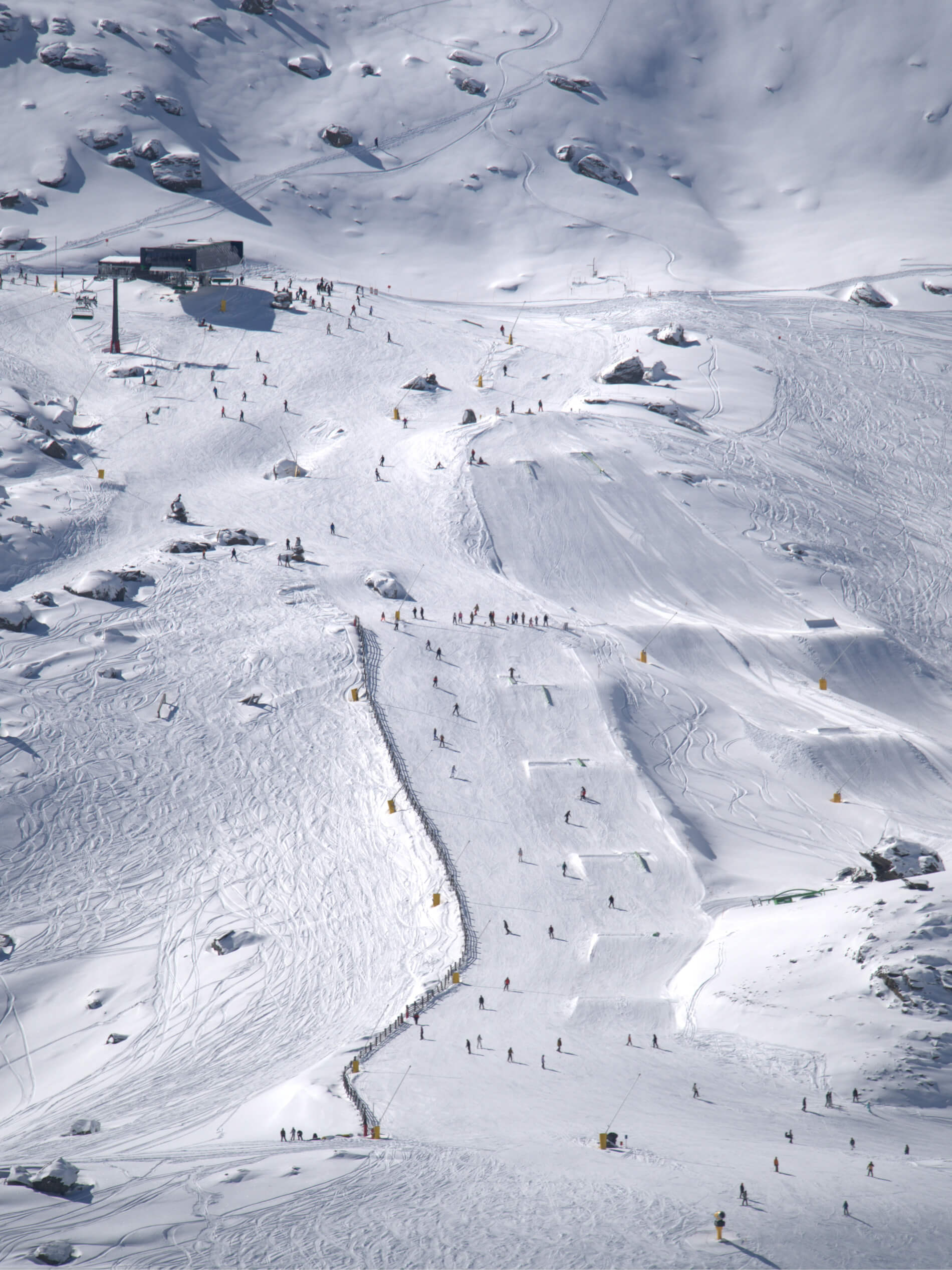 Wide view of a snowy ski slope with skiers and snowboarders near ski lift and rocks.