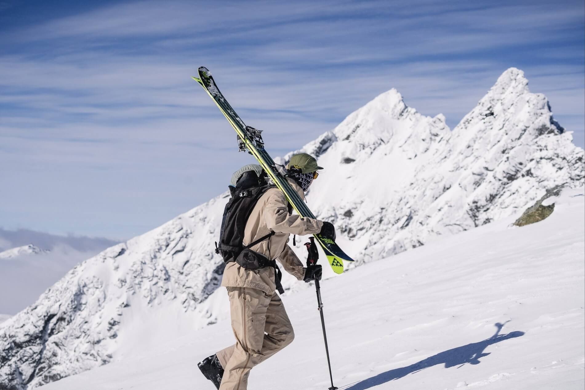 Skier hiking uphill in snow carrying skis on shoulder with snowy mountain peaks in the background.