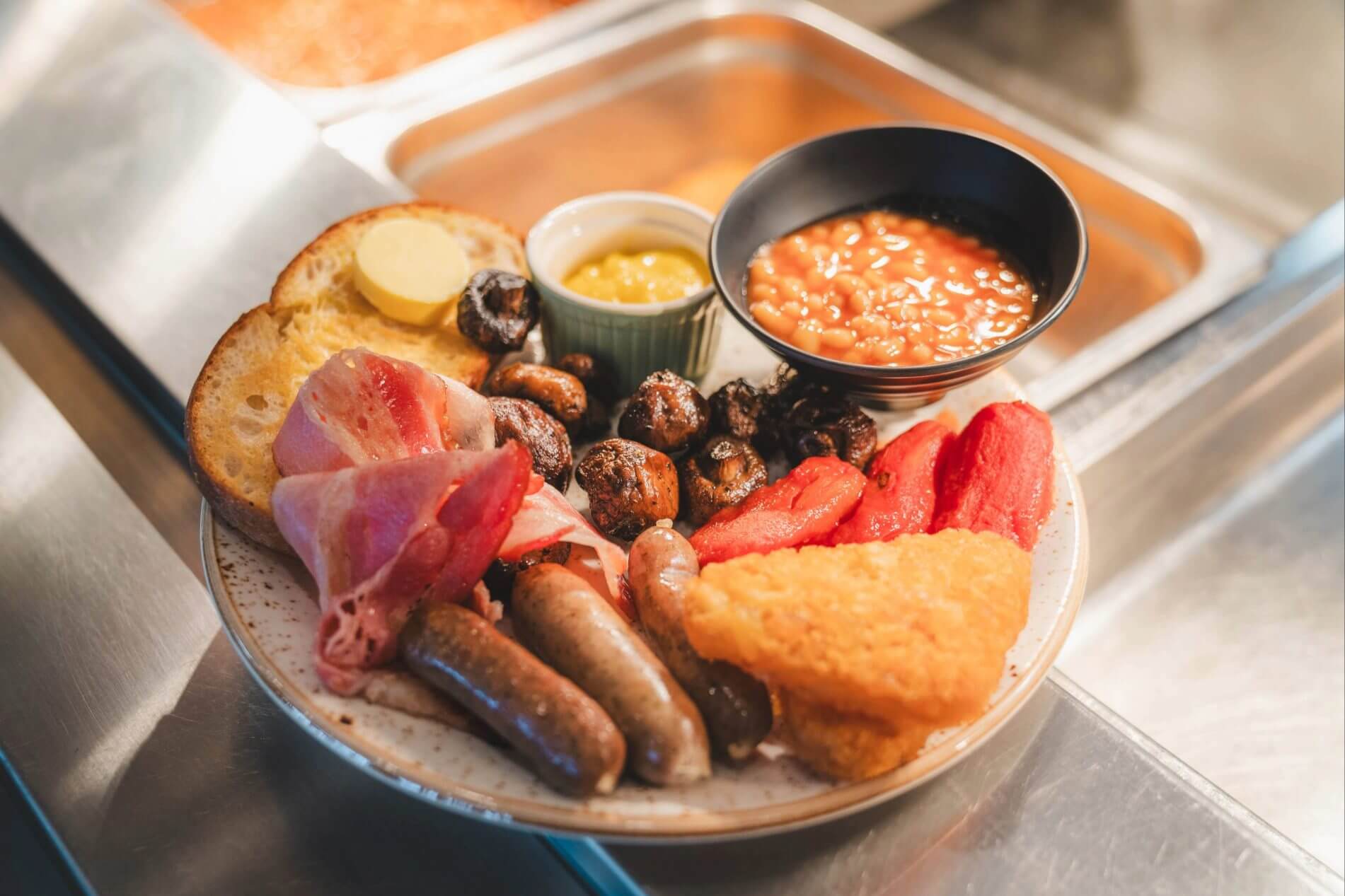 Full breakfast plate with sausages, bacon, mushrooms, baked beans, grilled tomatoes, hash brown, toast, and butter.