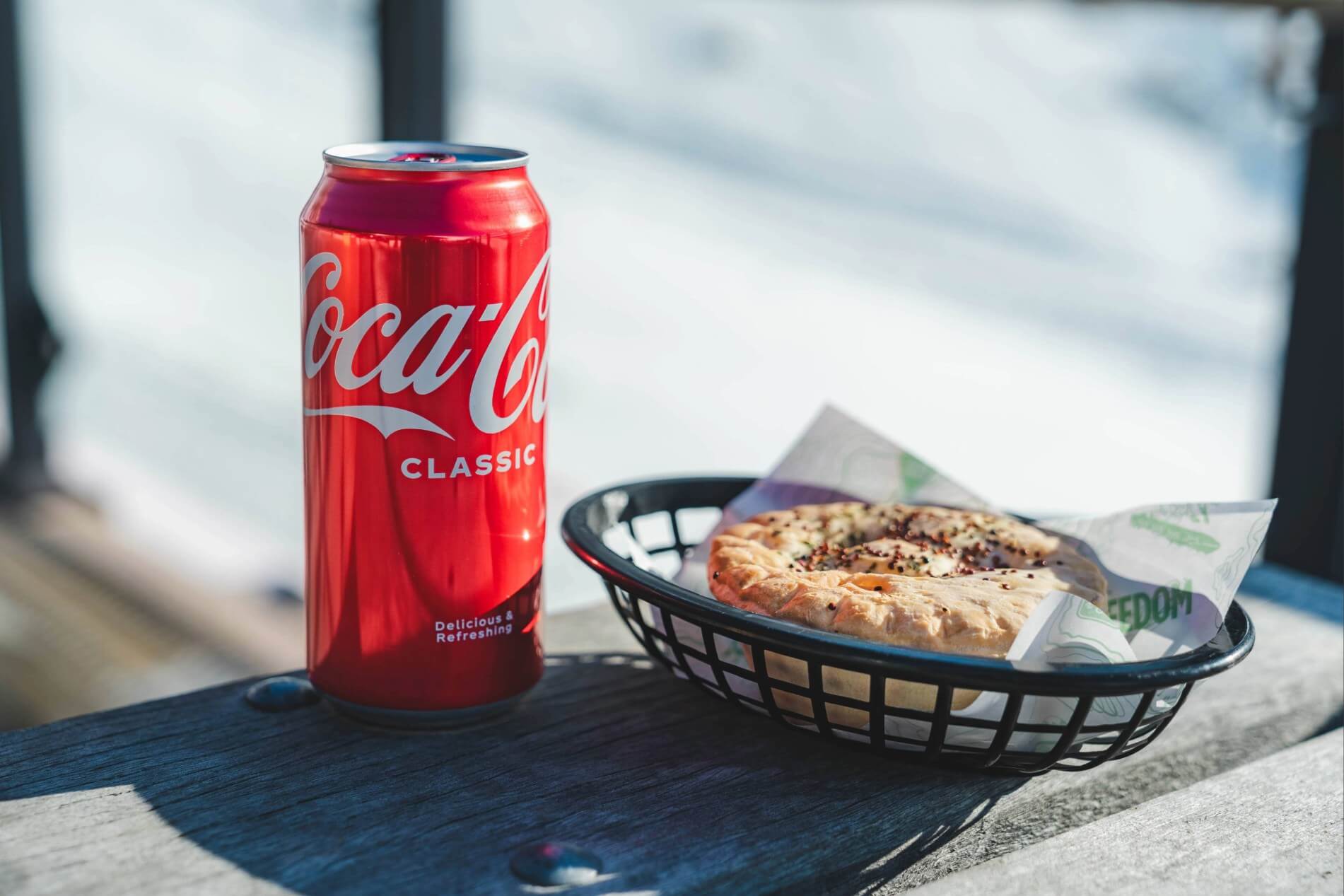 Red Coca-Cola Classic can beside a black basket holding a round, seasoned bread on a wooden table.