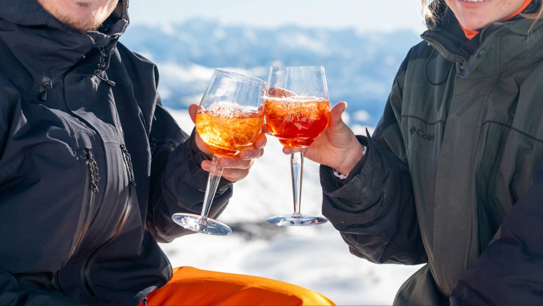 Two people in jackets clinking glasses of orange spritz drinks with snowy mountains background.
