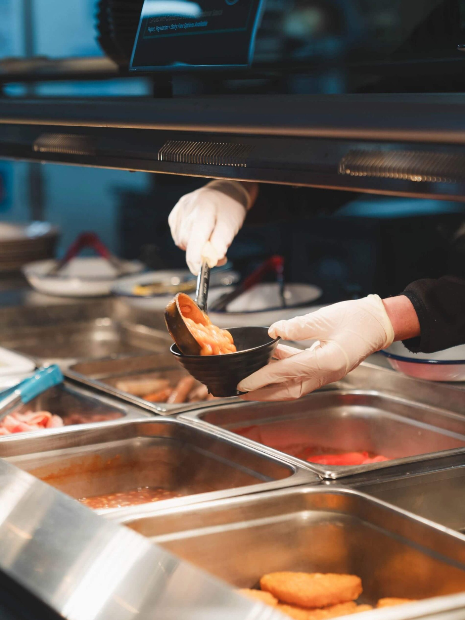 Gloved hands ladle hot food into a black bowl at a serving counter with metal trays.