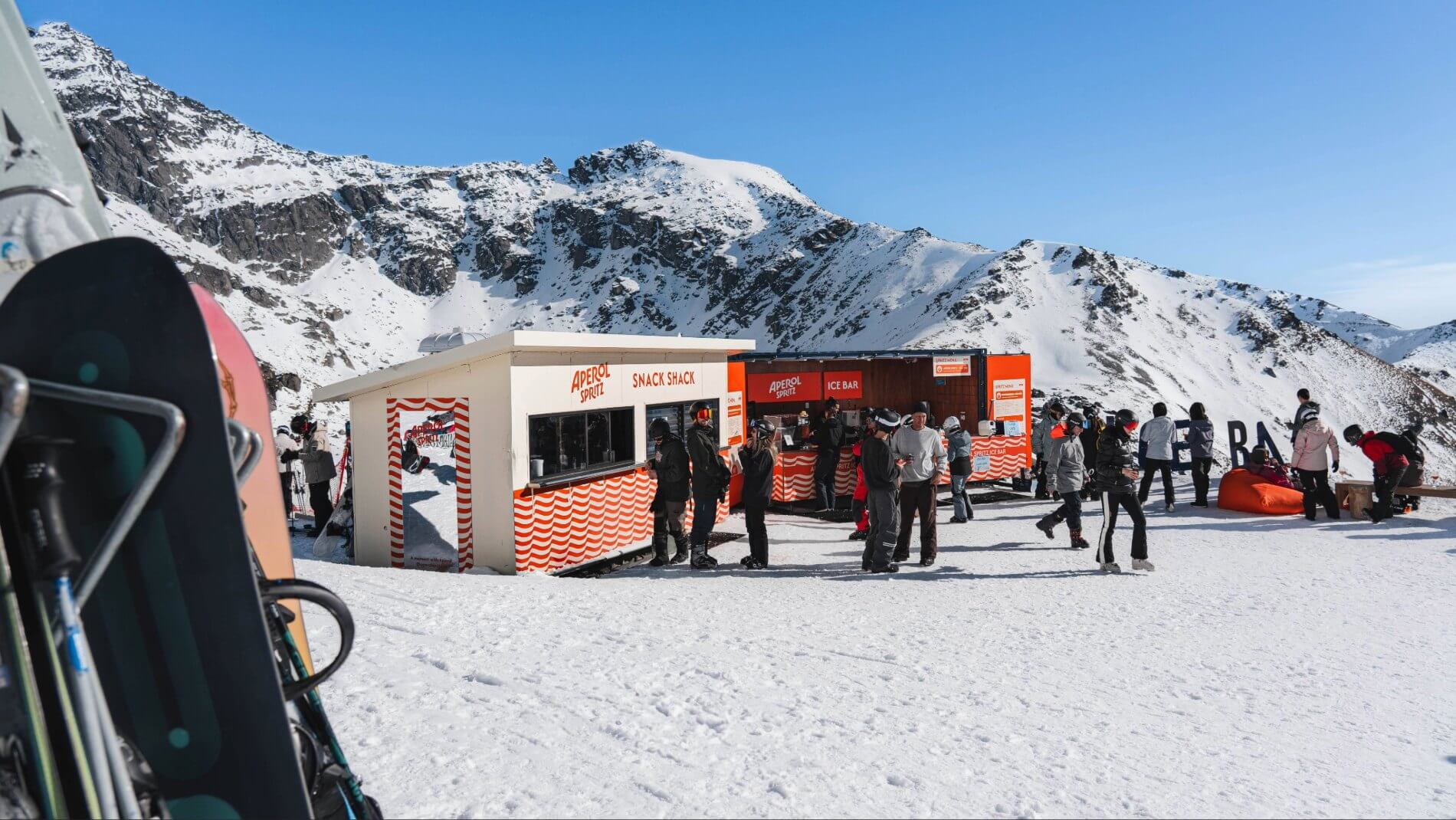People gathered around a snack shack and ice bar on a sunny snowy mountain with skis in foreground.