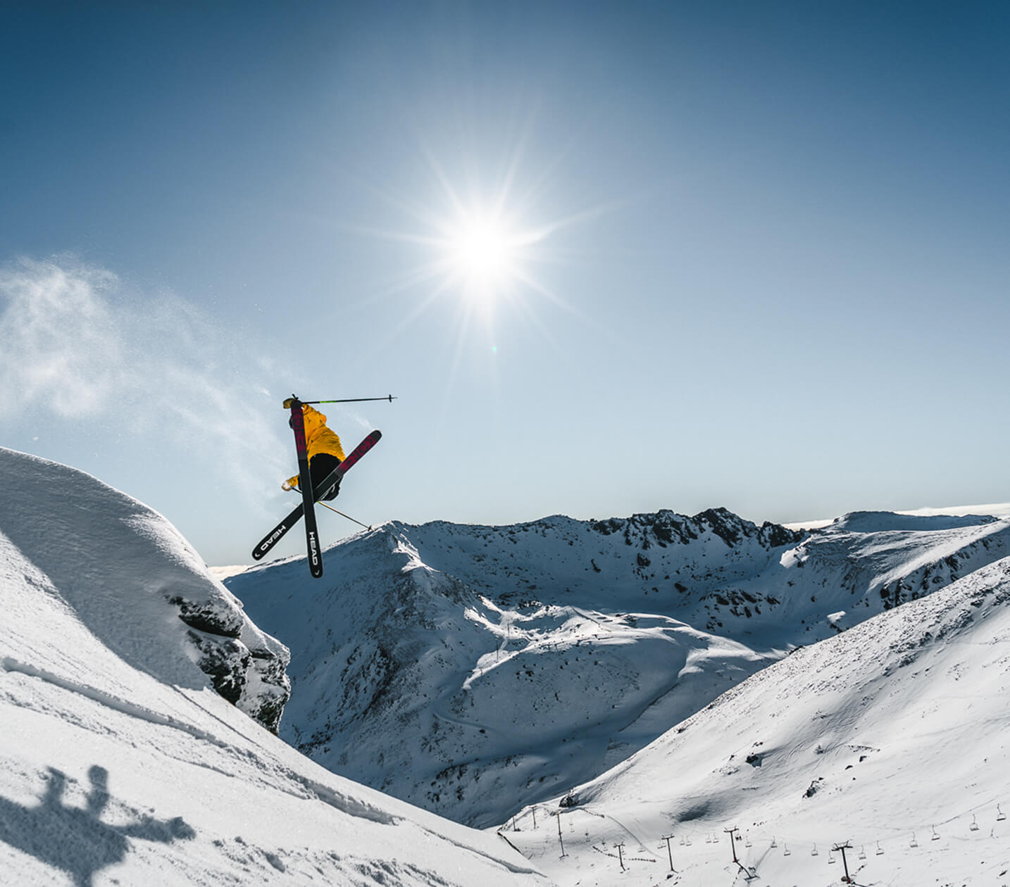 Skier in yellow jacket performing a jump over snowy mountain slopes under a bright sun.