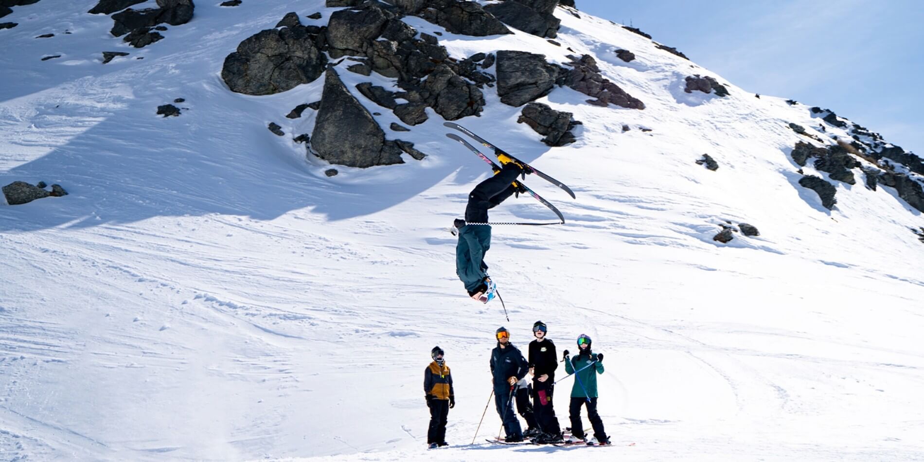 Skier doing a backflip in midair above four people standing on snow near rocky mountain slope.