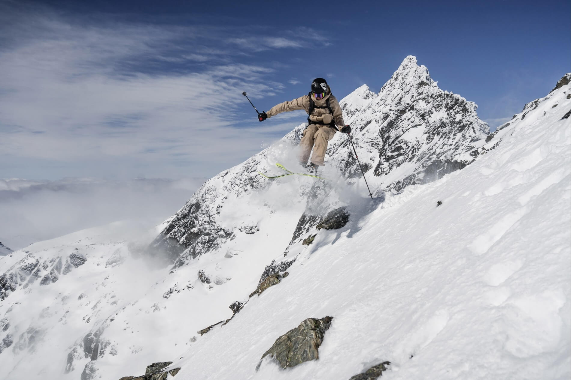 Skier in beige gear jumps off a snowy slope with rugged mountain peaks in the background.