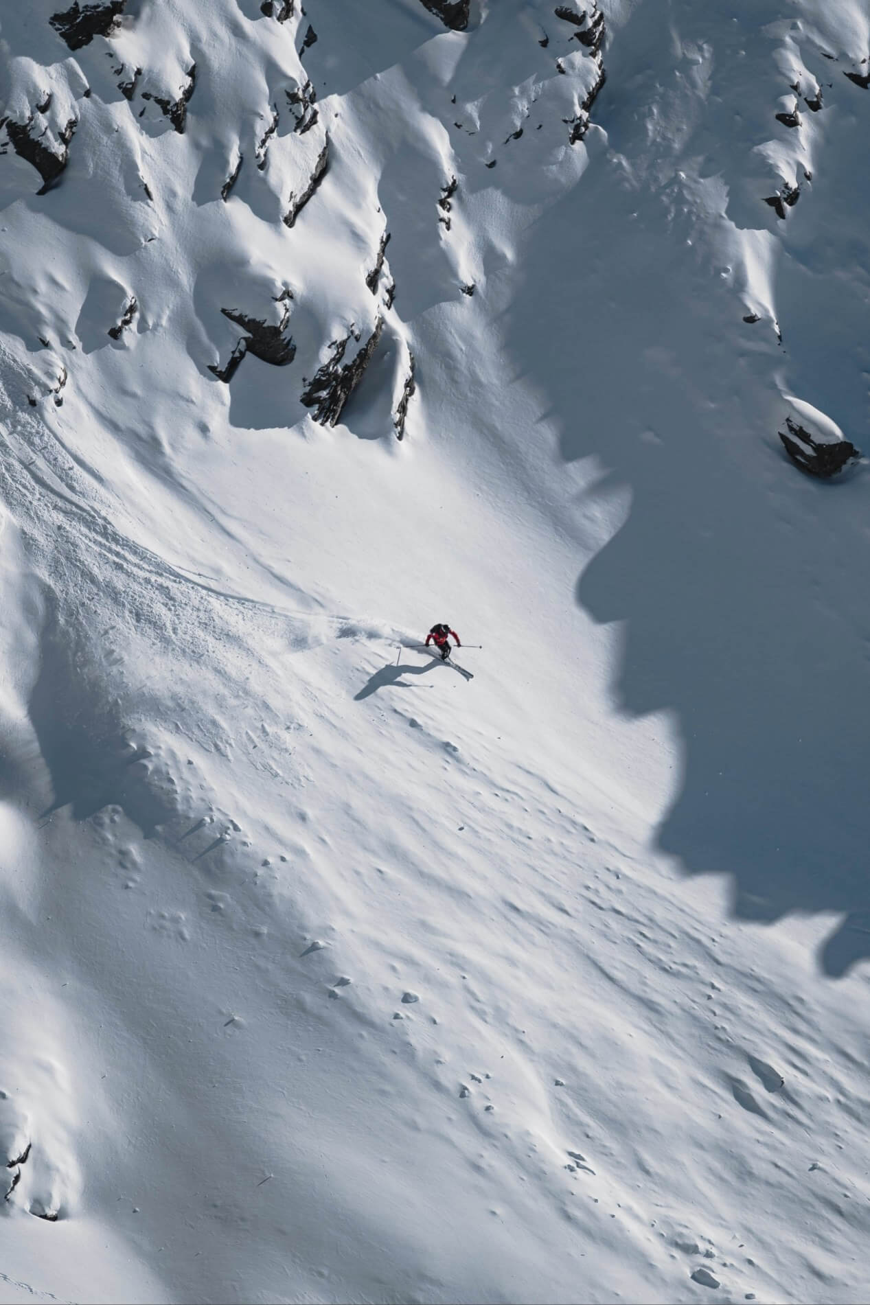 A lone skier making a turn down a snowy mountain face with rocky outcrops.