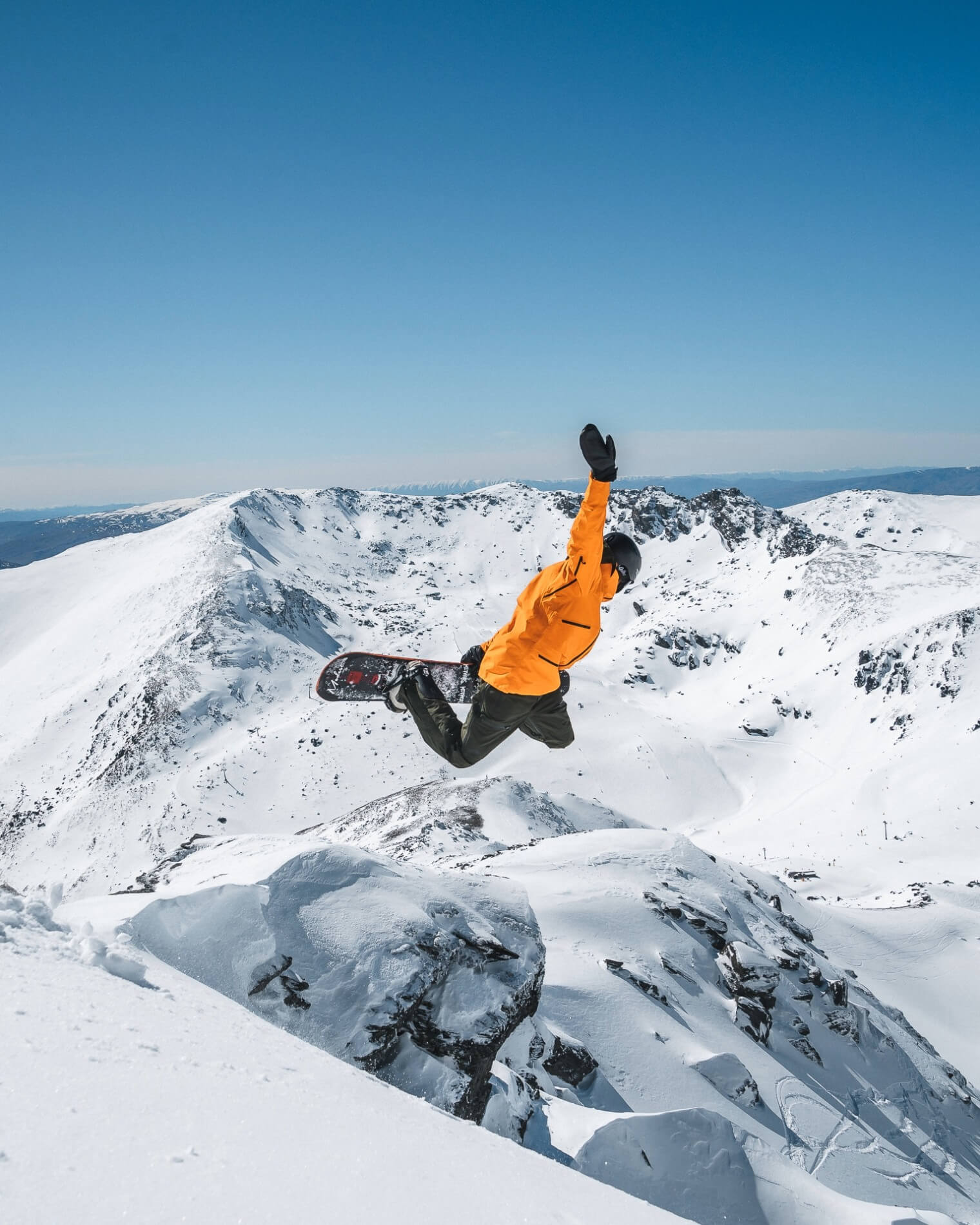 Snowboarder in orange jacket performing a jump grab in a snowy mountain landscape under a clear blue sky.