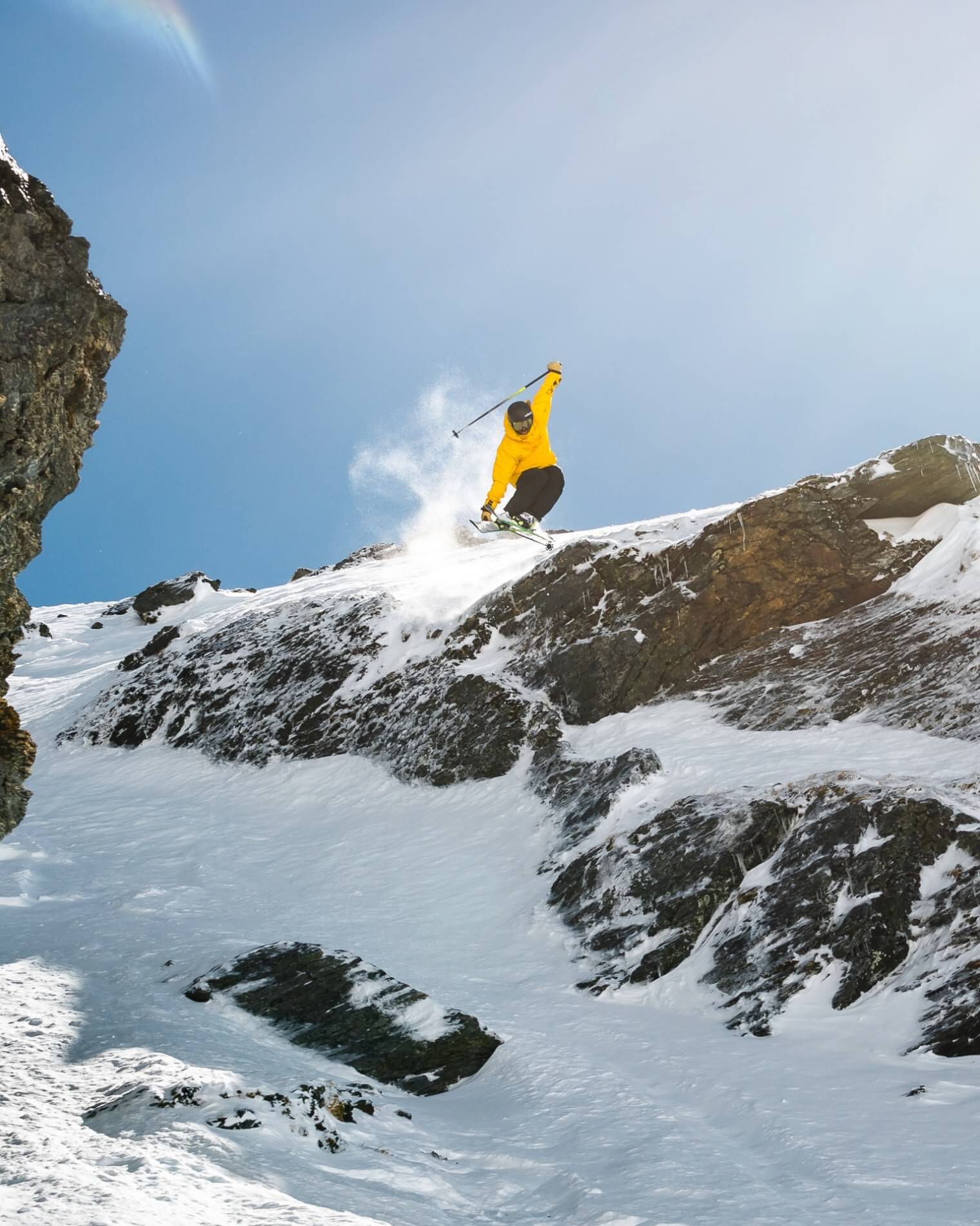 Skier in yellow jacket jumping off a snowy rocky mountain cliff under a clear blue sky.