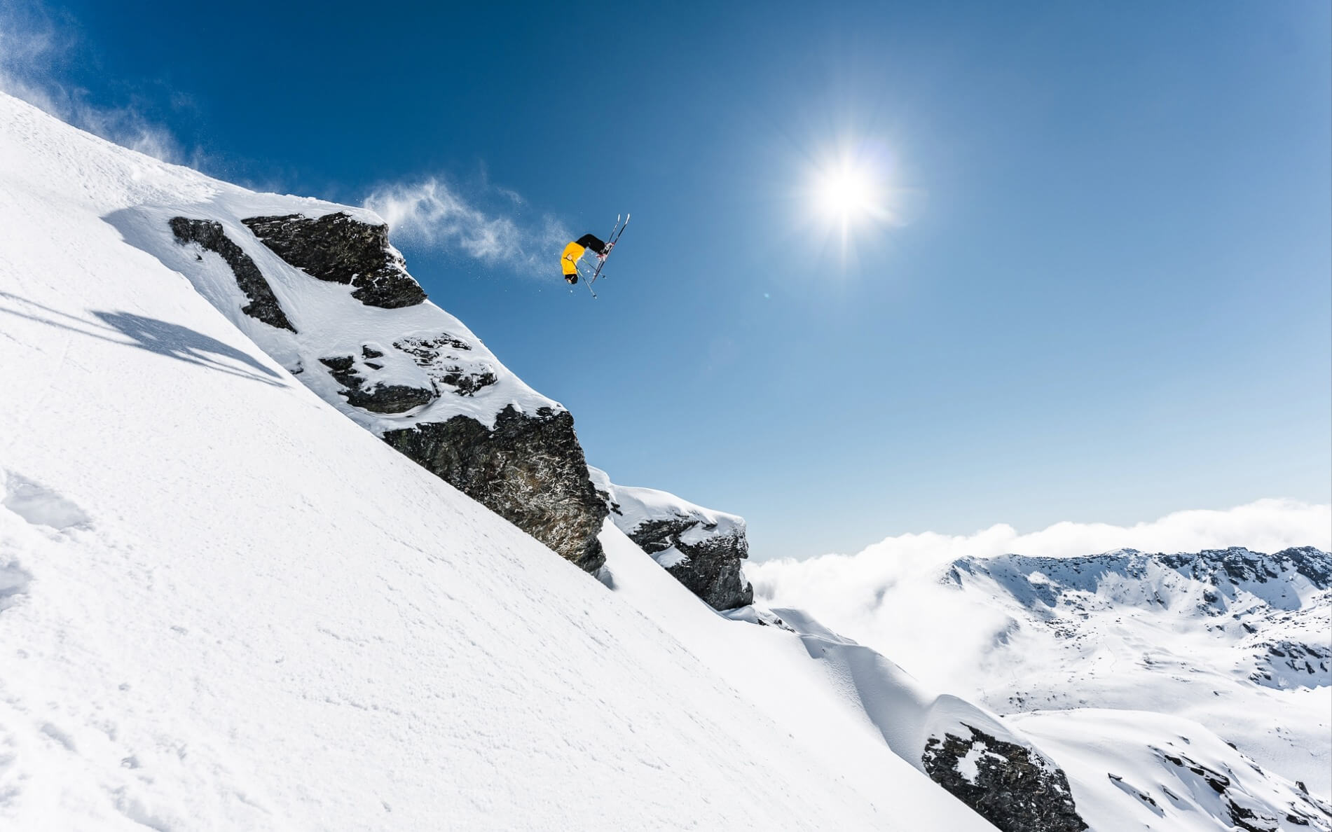Skier in yellow jacket performing a backflip off a snowy mountain cliff under bright sun.