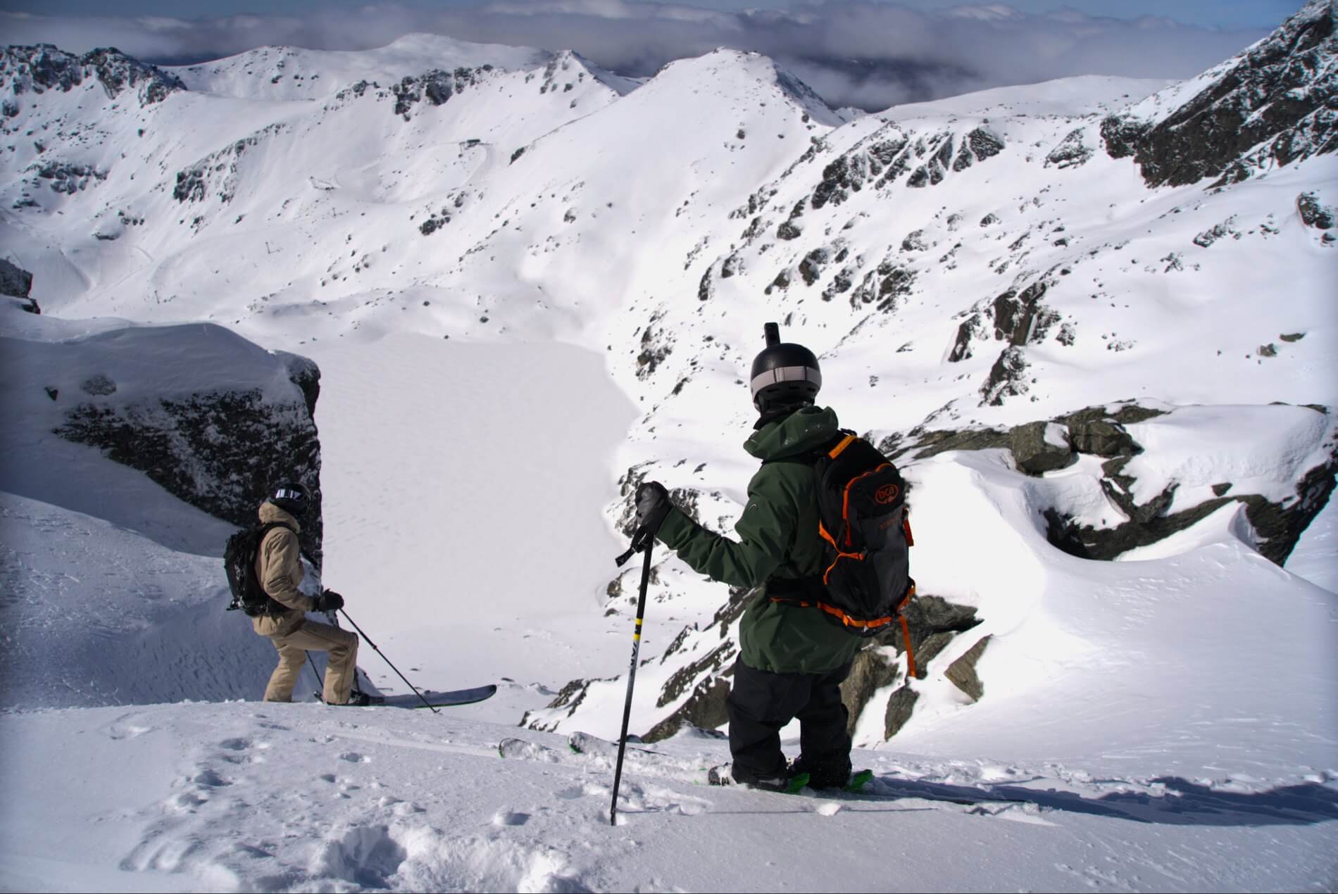 Two skiers in winter gear standing on a snowy mountain ridge overlooking snow-covered peaks.
