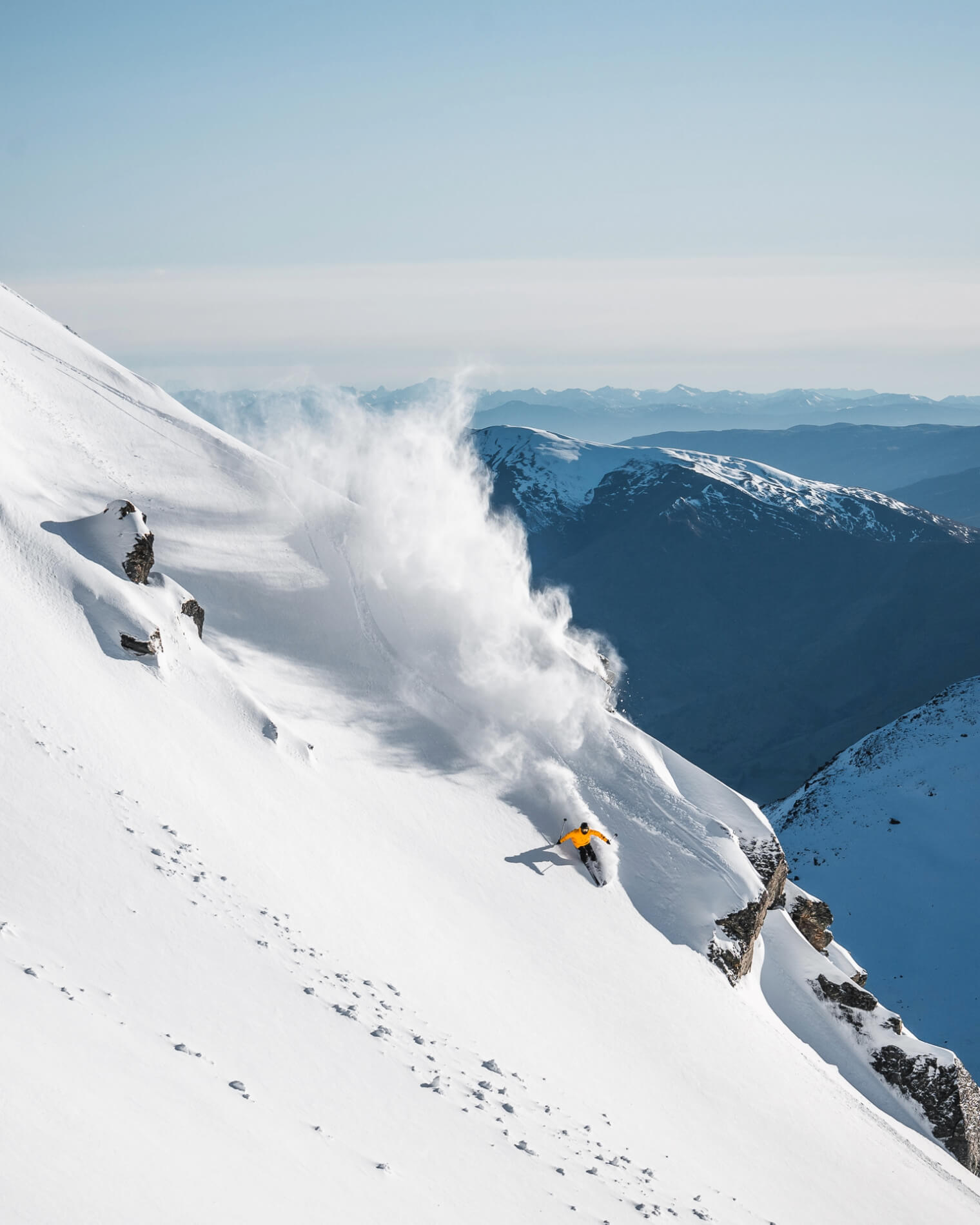 Skier in a yellow jacket skiing down a snowy mountain with snow spray and mountain range background.