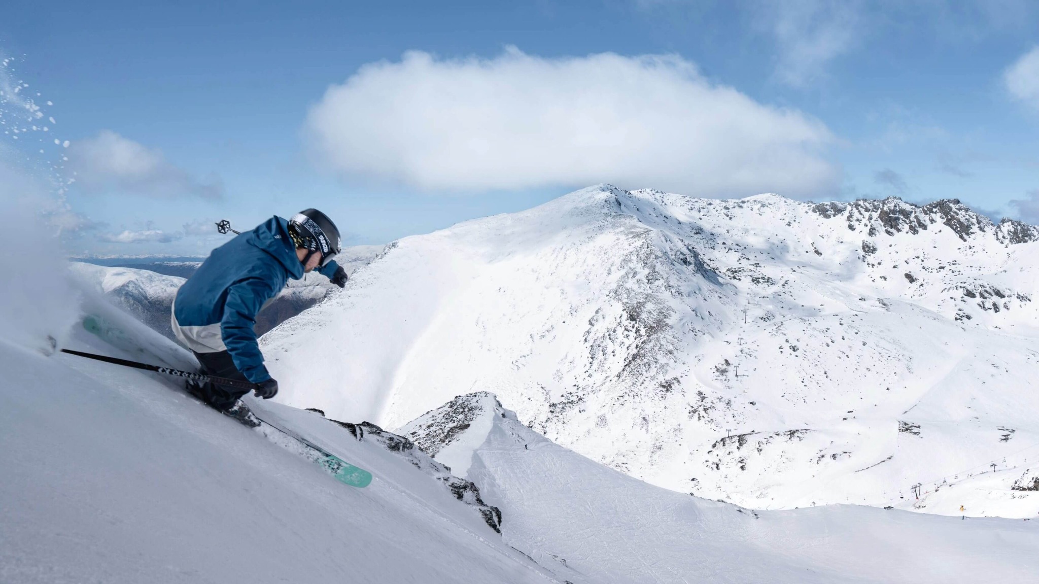 Skier in a blue jacket carving down a steep snow-covered mountain slope under a blue sky.