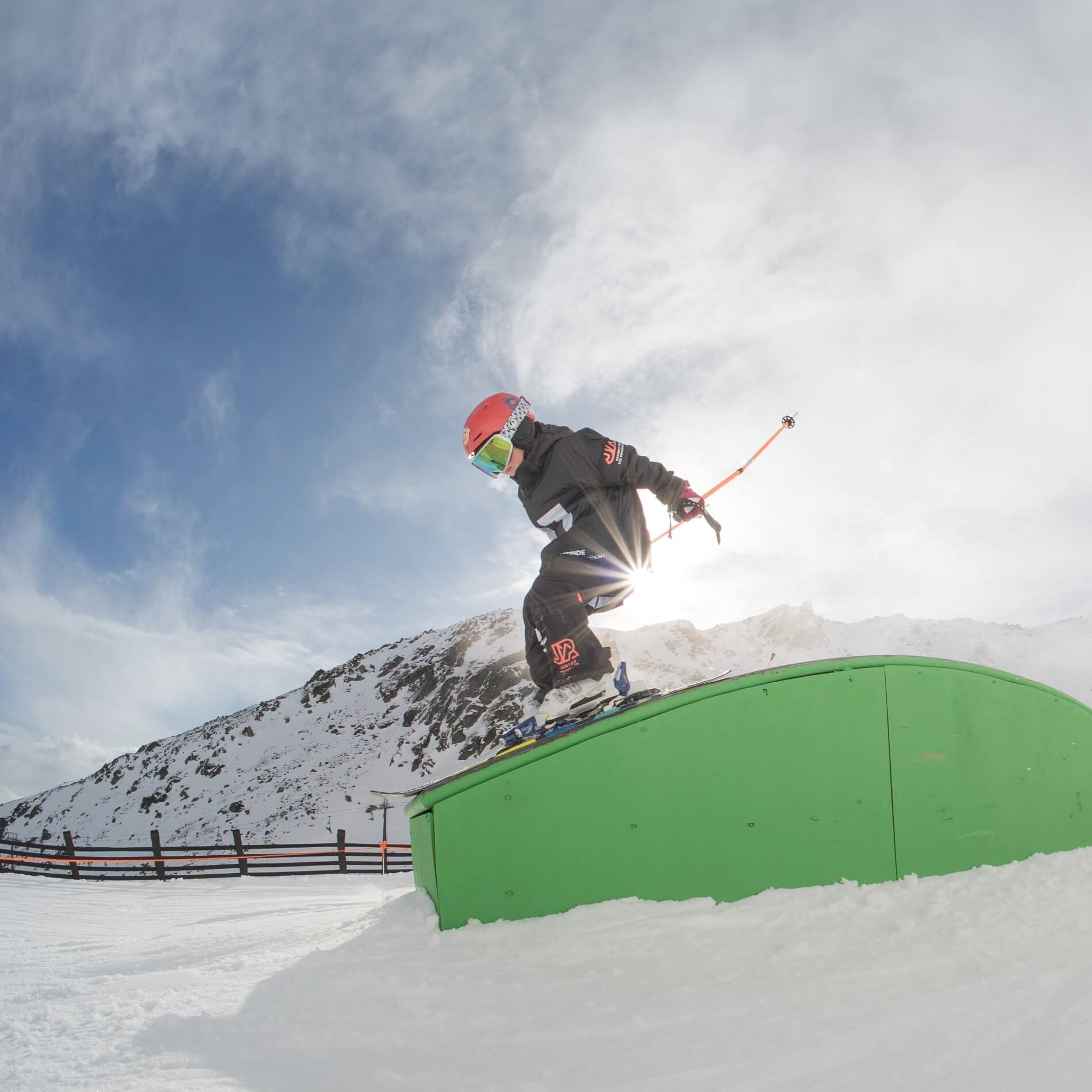 Child skier in red helmet sliding on a green rail in a snowy mountain terrain park.