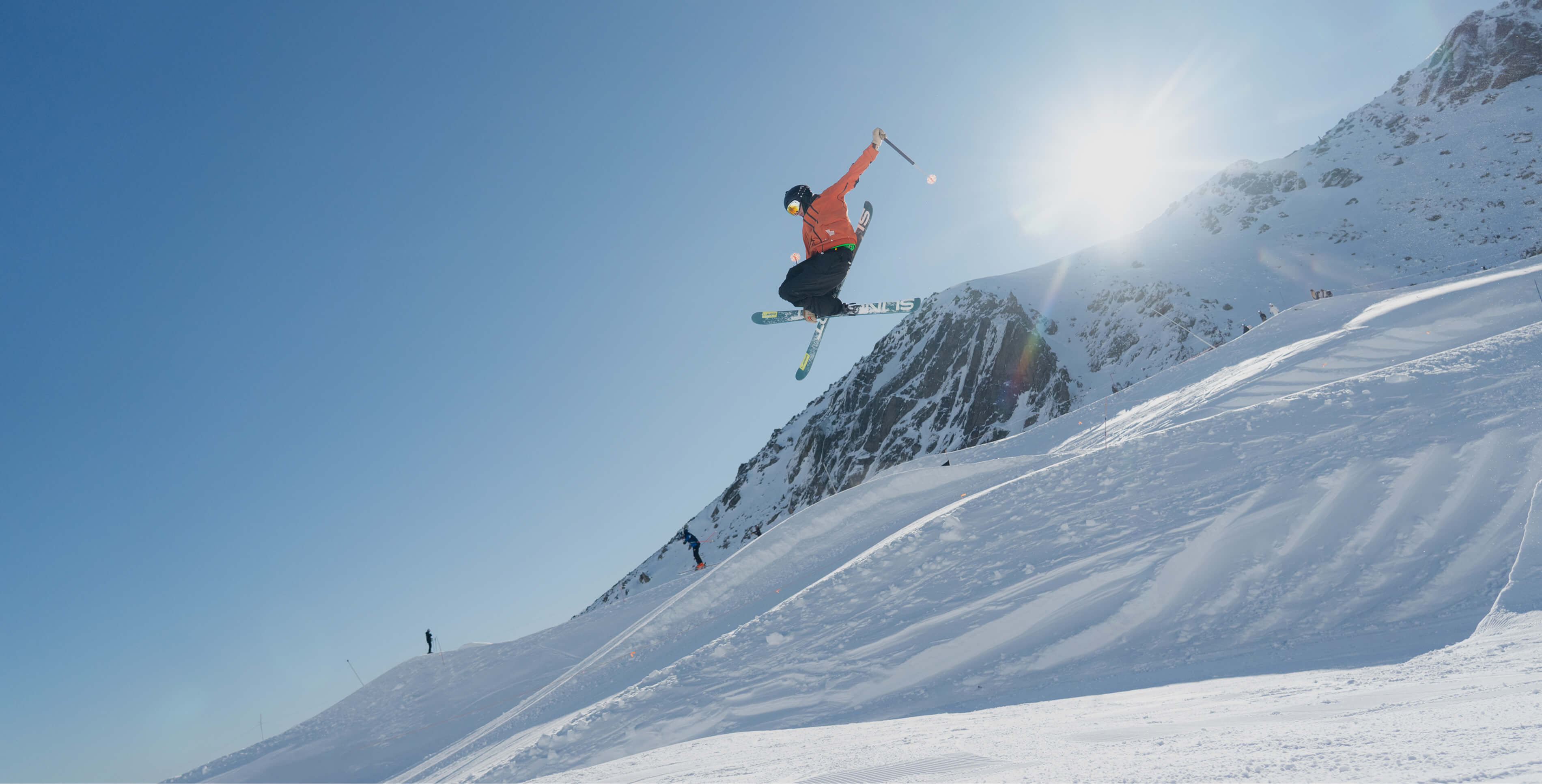 Skier in an orange jacket performs a high jump with poles on a snowy mountain under bright sun.