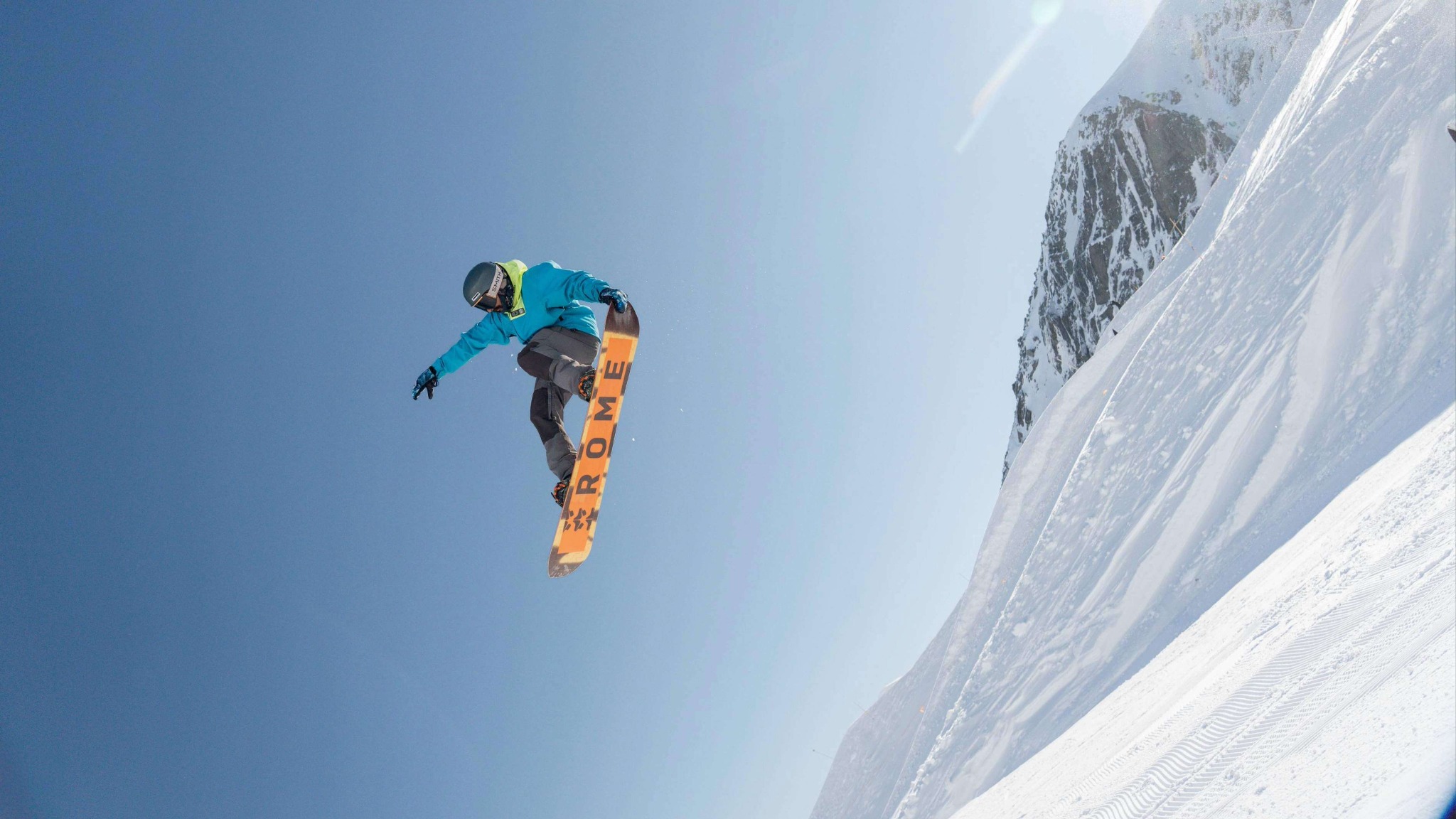 Snowboarder in blue jacket performing a high jump against clear blue sky over snowy mountain.