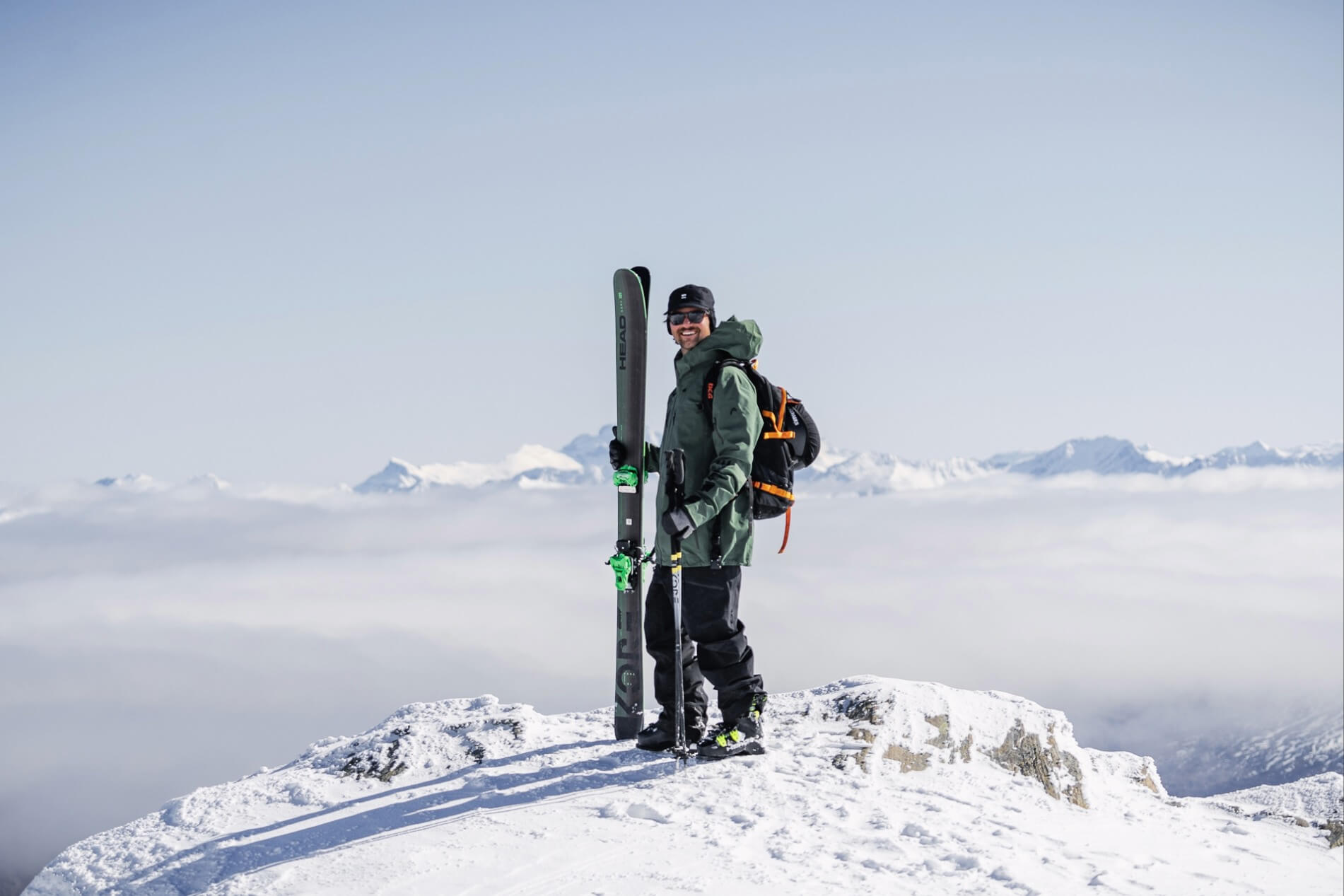 Smiling skier in green jacket holding skis atop snowy mountain with peaks in the distance.