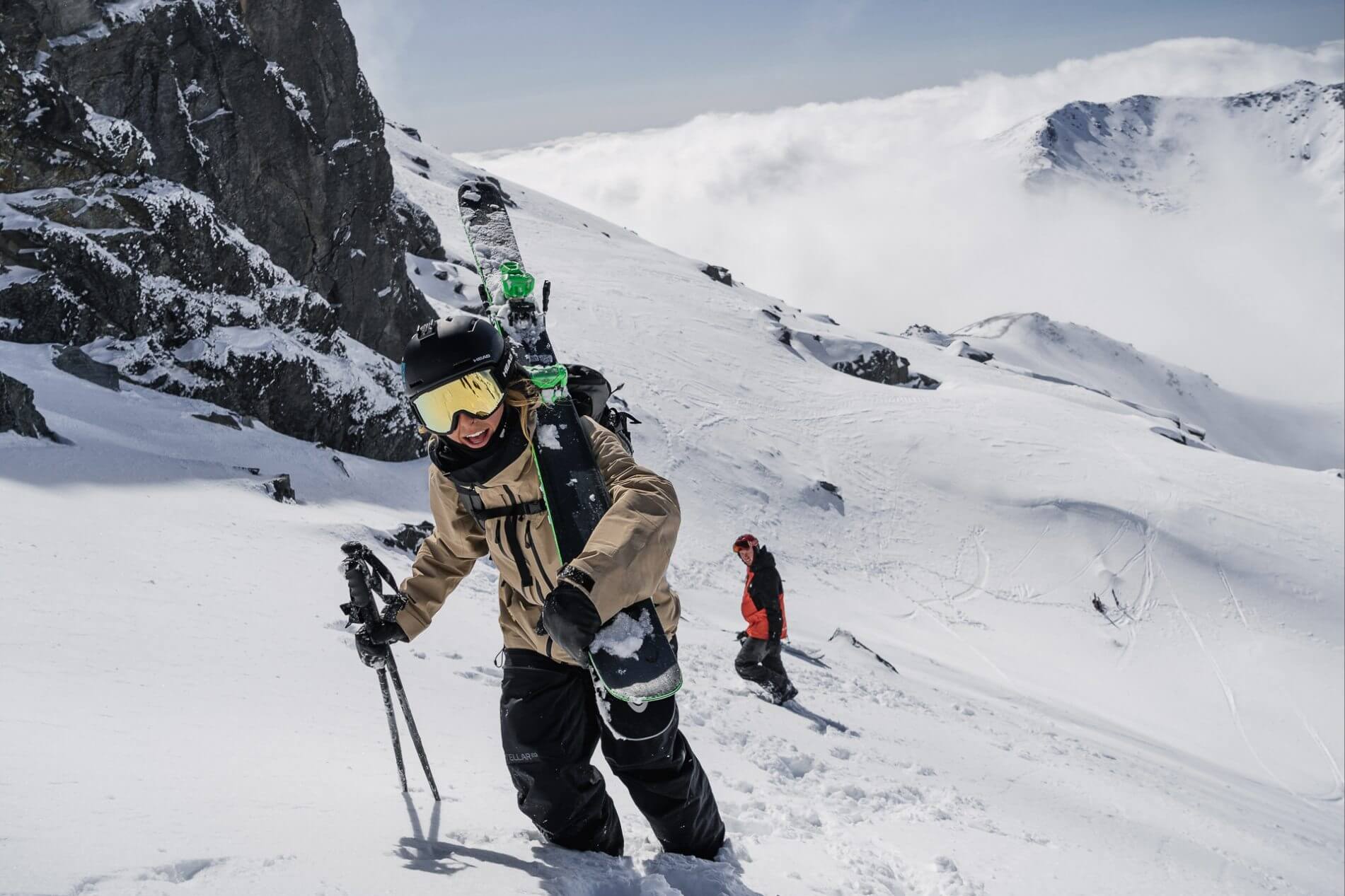 Backcountry skier carrying skis and poles climbs snowy mountain slope with cloudy peaks behind.