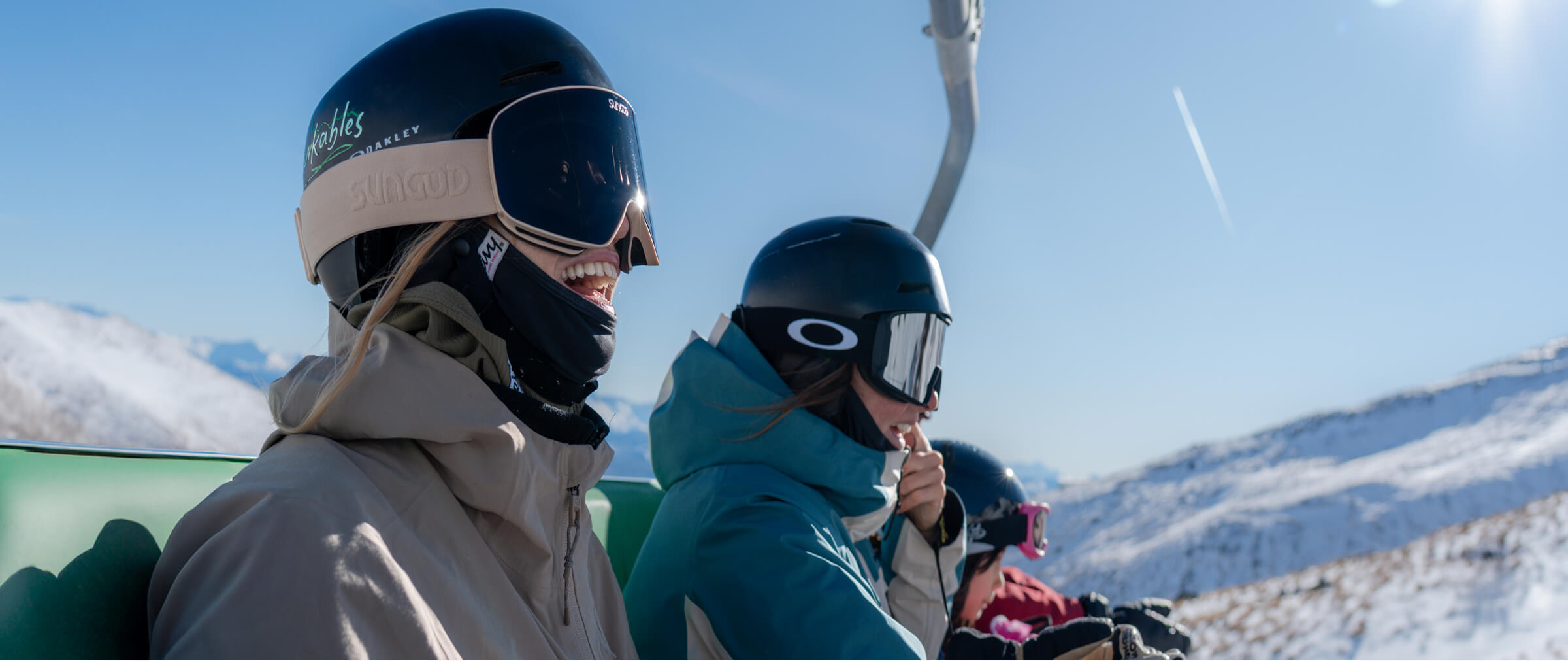 Three skiers in helmets and goggles riding a chairlift over snowy mountains on a clear day.