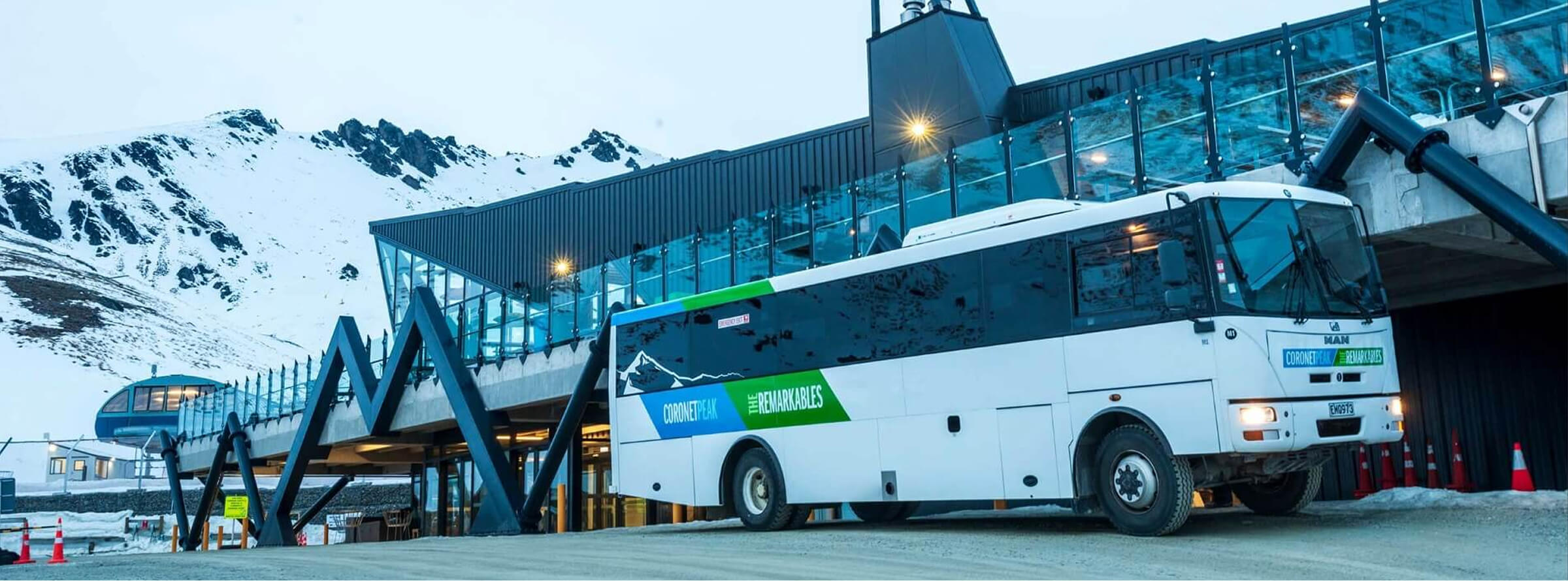 White shuttle bus with Coronet Peak and The Remarkables logo parked beside a mountain lodge under snowy peaks.