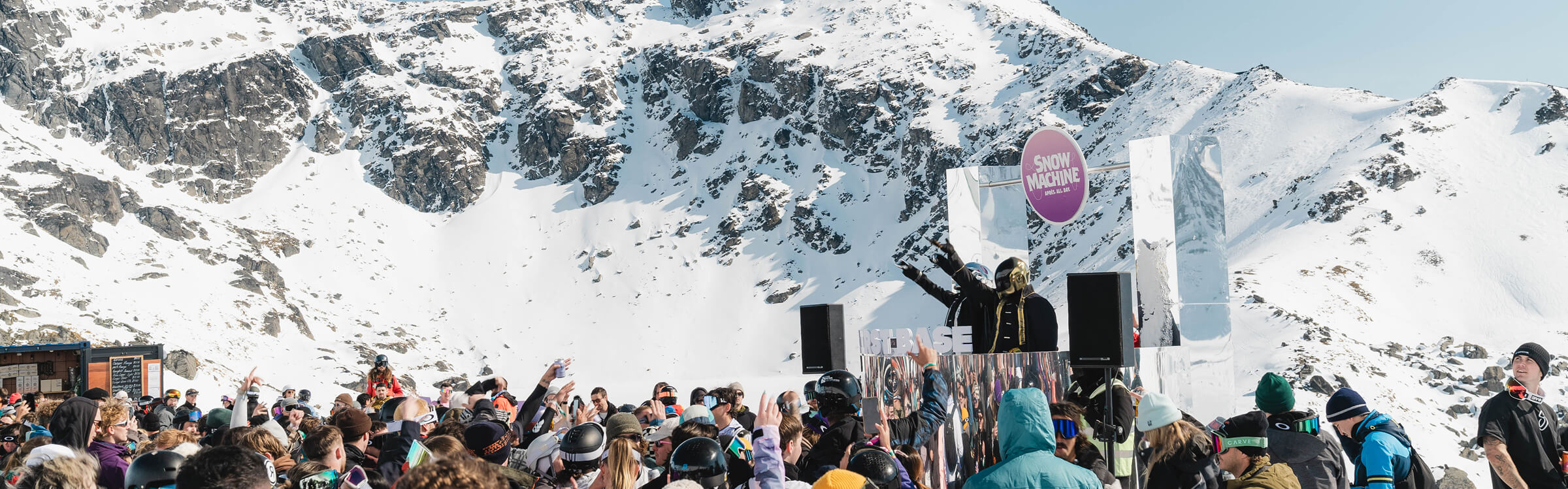Crowd in winter gear enjoying an outdoor music event with snow-covered mountains in the background.