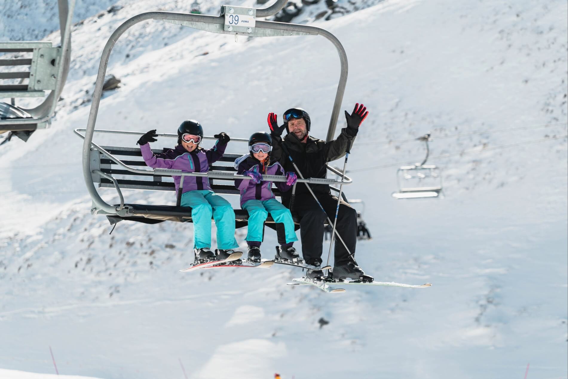 Man and two children in ski gear smiling and waving from a ski chairlift above snow.