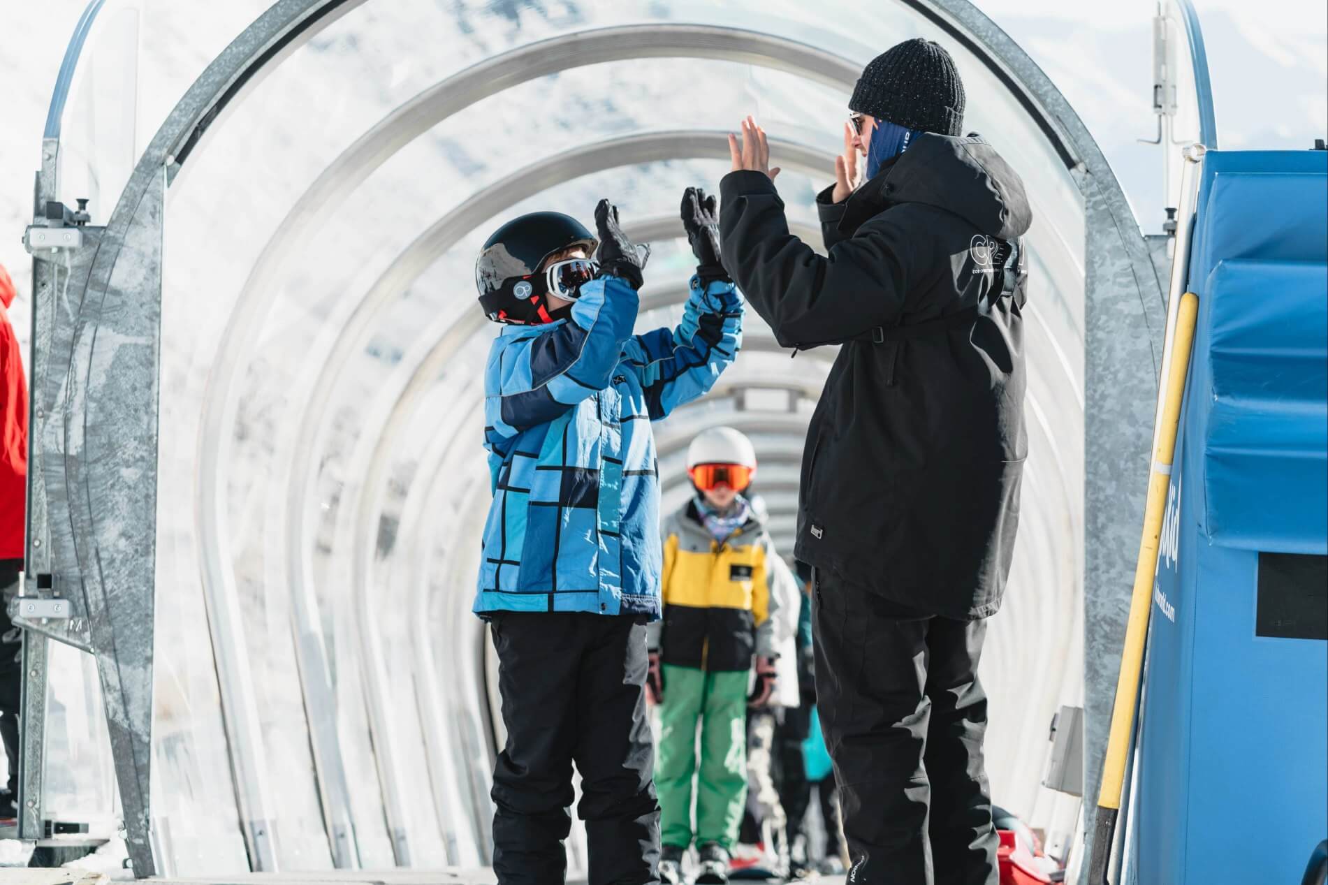 Child and adult in ski gear giving high five inside a tubular snow conveyor belt.