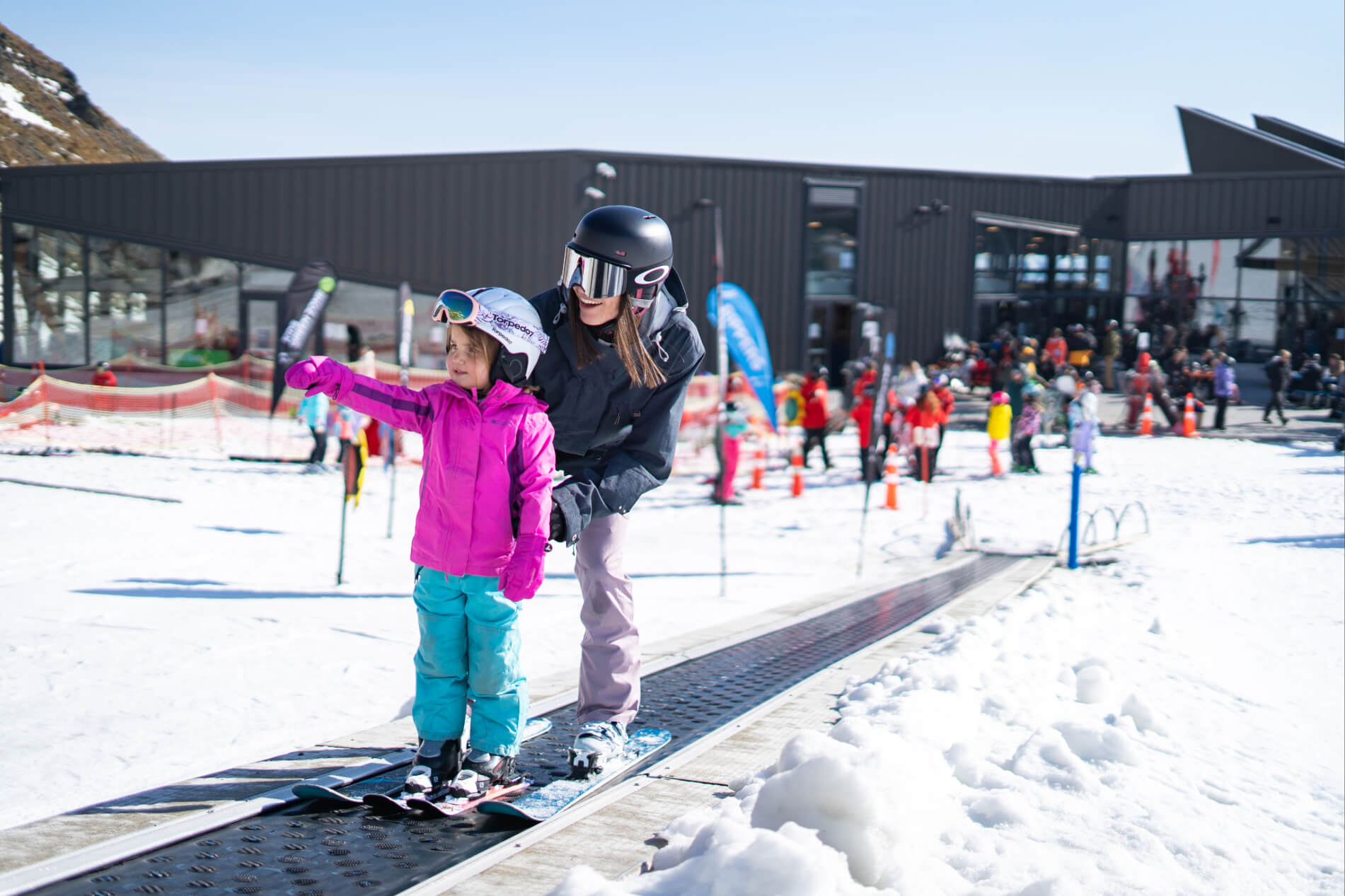 Child in pink jacket and helmet learning to ski with adult instructor on snow near ski lodge.