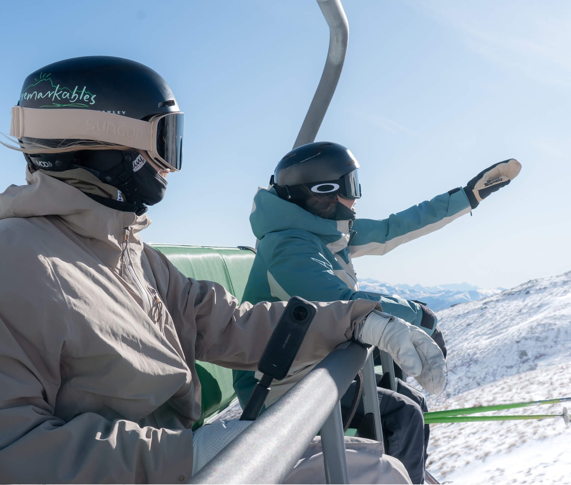 Two skiers wearing helmets and goggles sitting on a ski lift with snowy mountains in the background.