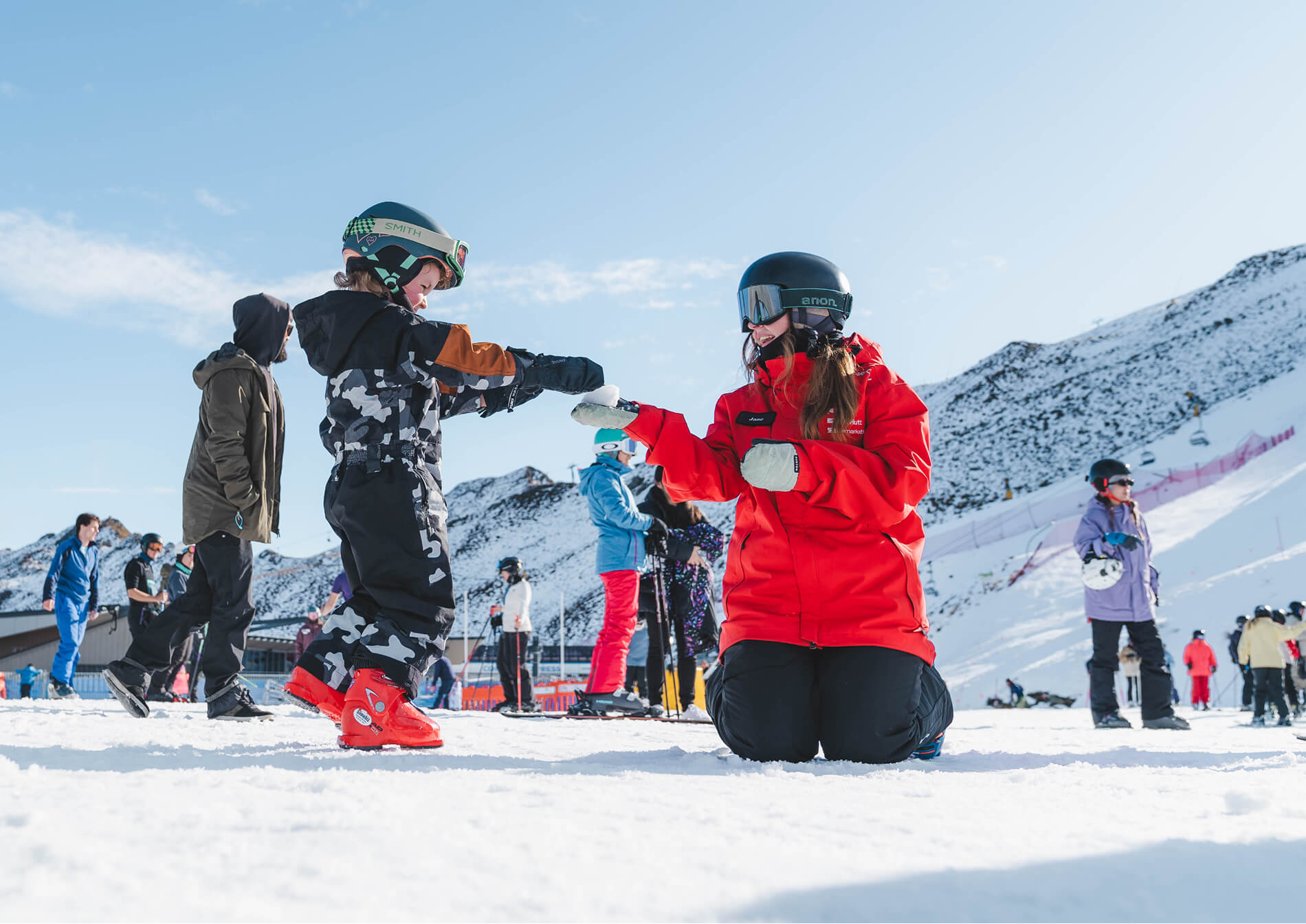 Child in ski gear handing snow to kneeling ski instructor in red jacket on snowy slope with mountains.