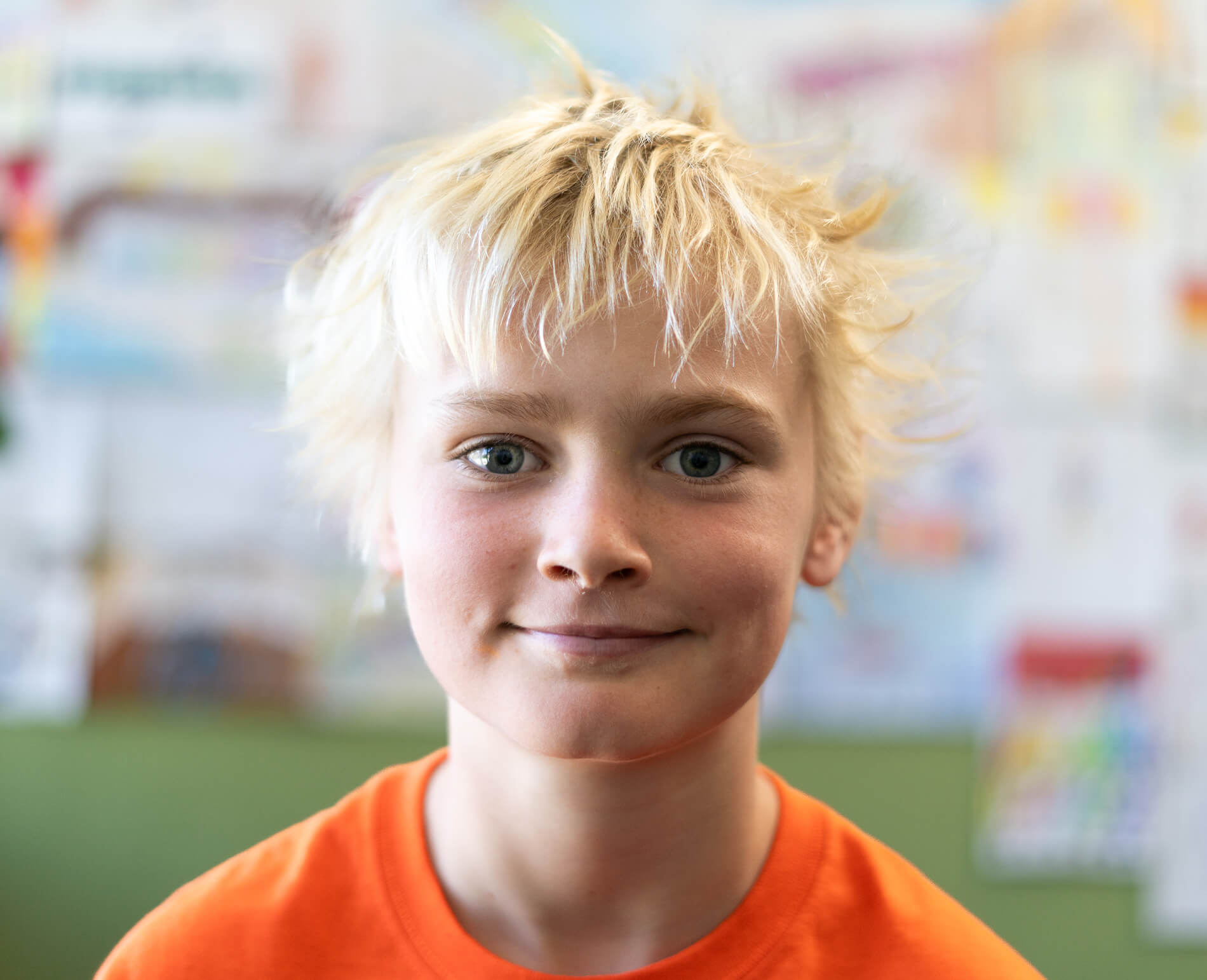 Smiling blonde boy with tousled hair wearing an orange shirt in a softly blurred room.