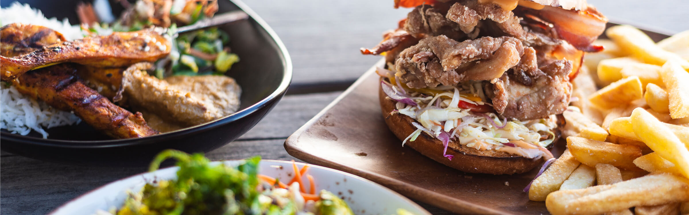 Fried chicken sandwich with coleslaw and fries on a wooden tray, with grilled chicken and rice dish nearby.