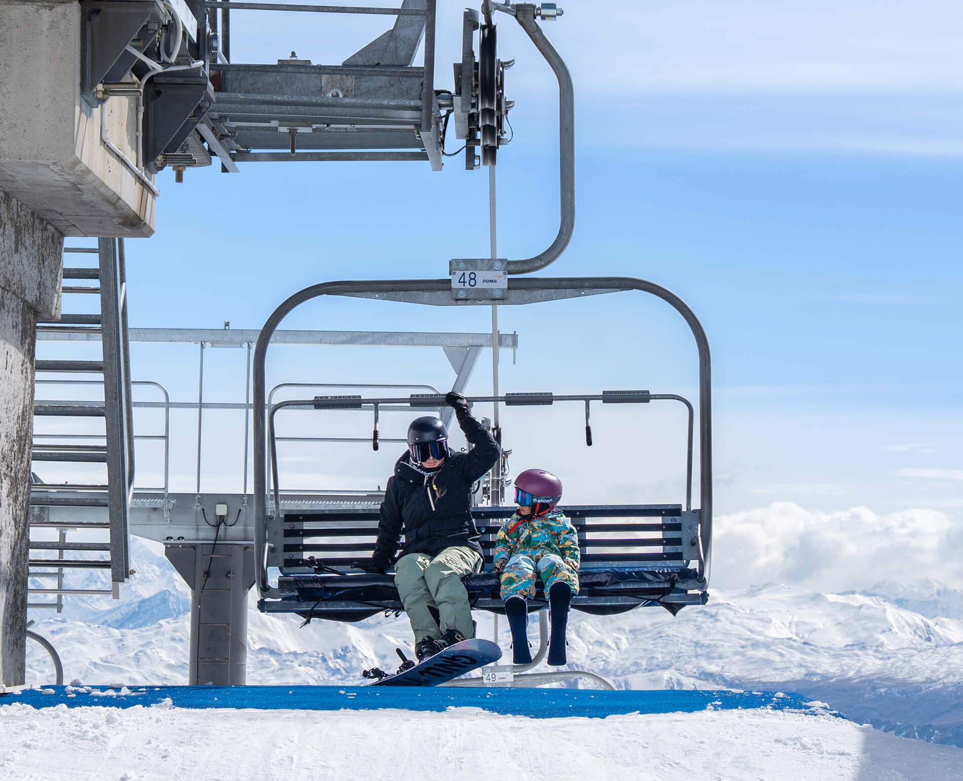 Person and child sitting on ski lift in snowy mountain landscape wearing helmets and winter gear.