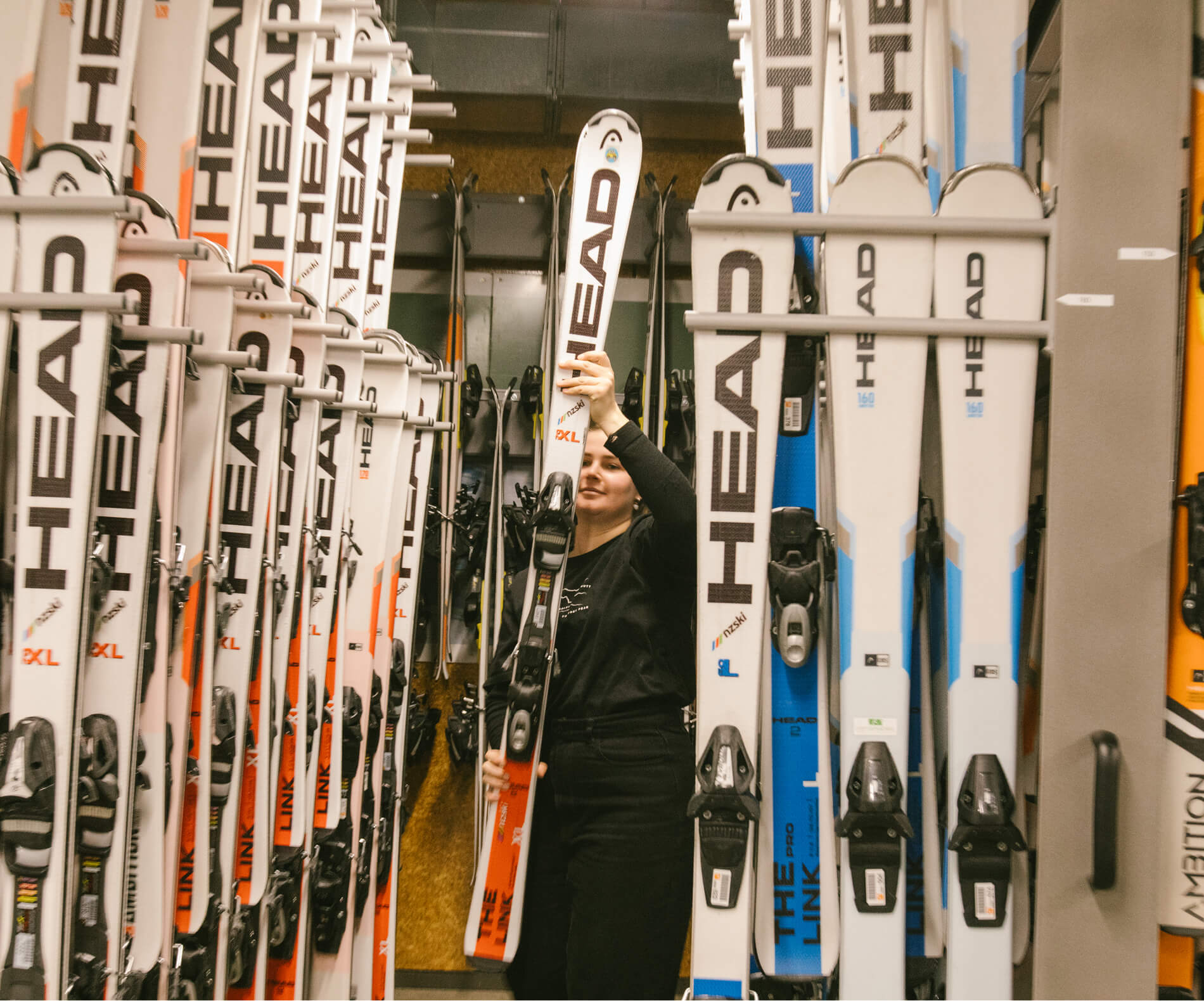 Person holding a pair of white HEAD skis in a ski rental or storage room filled with skis.