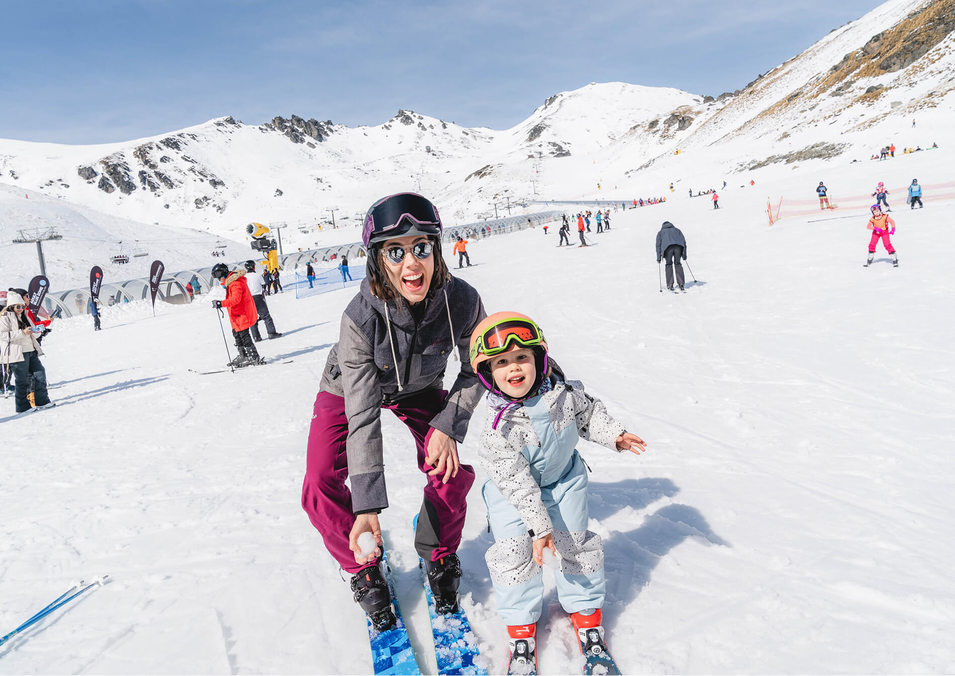 Adult and child smiling together while skiing on a snowy mountain slope with other skiers.