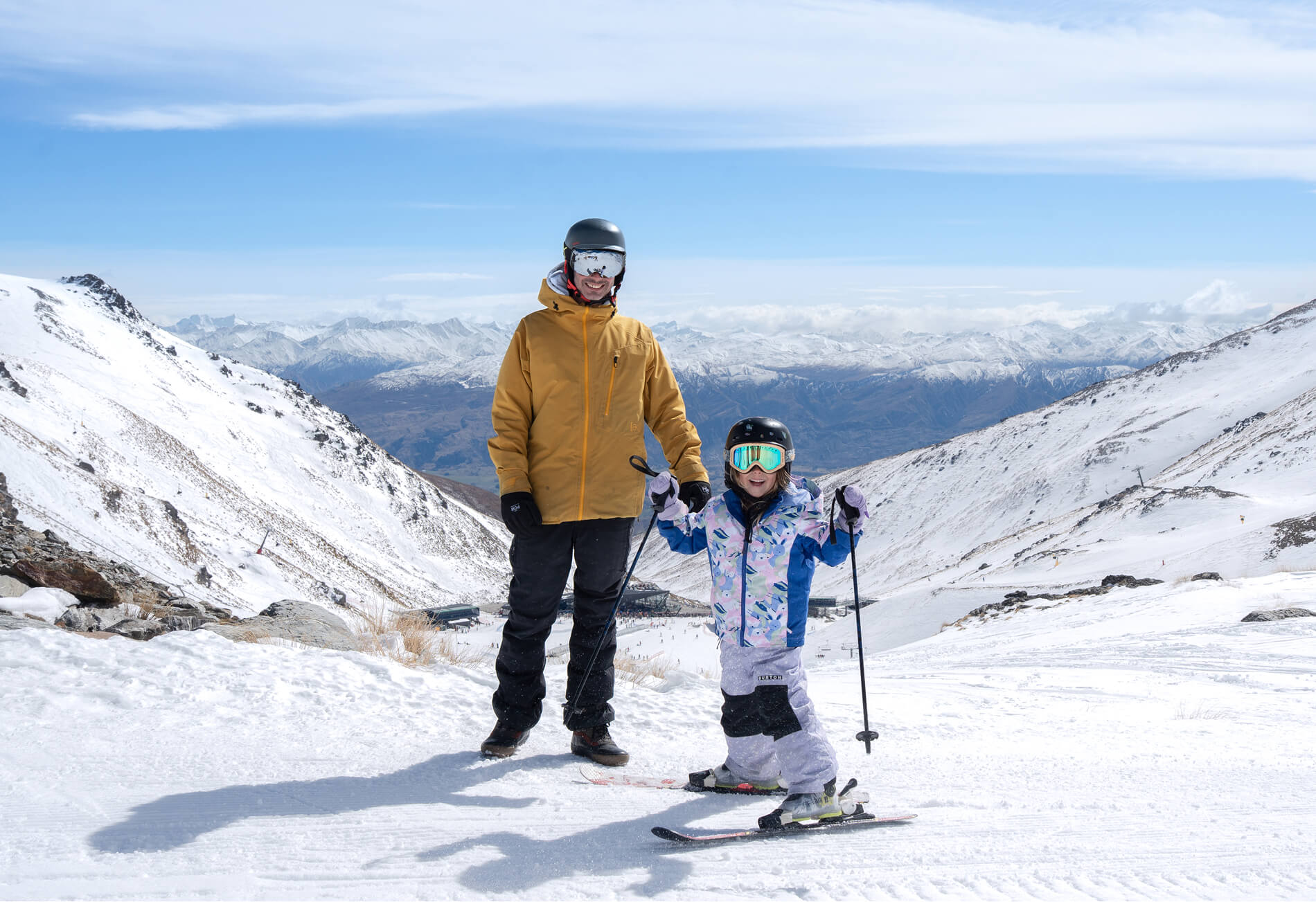Adult and child in ski gear standing on snowy mountain slope with ski poles and skis.
