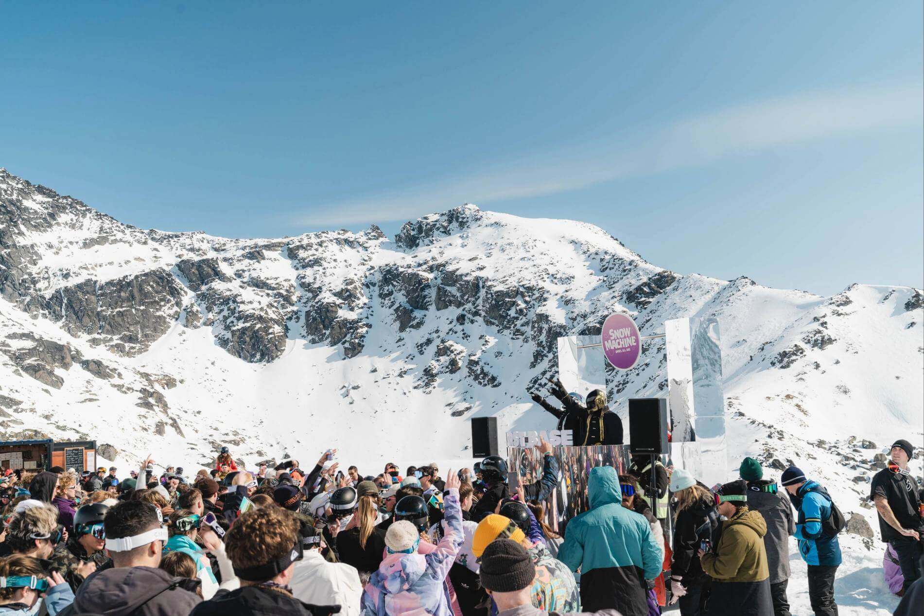 Crowd enjoying an outdoor DJ event in snowy mountains under clear blue sky.