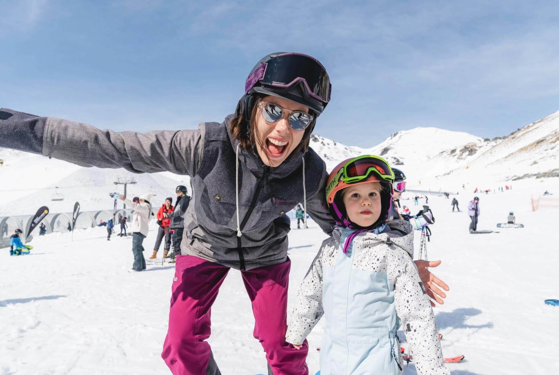 Smiling woman in ski gear poses with a child wearing a helmet on a snowy ski slope.