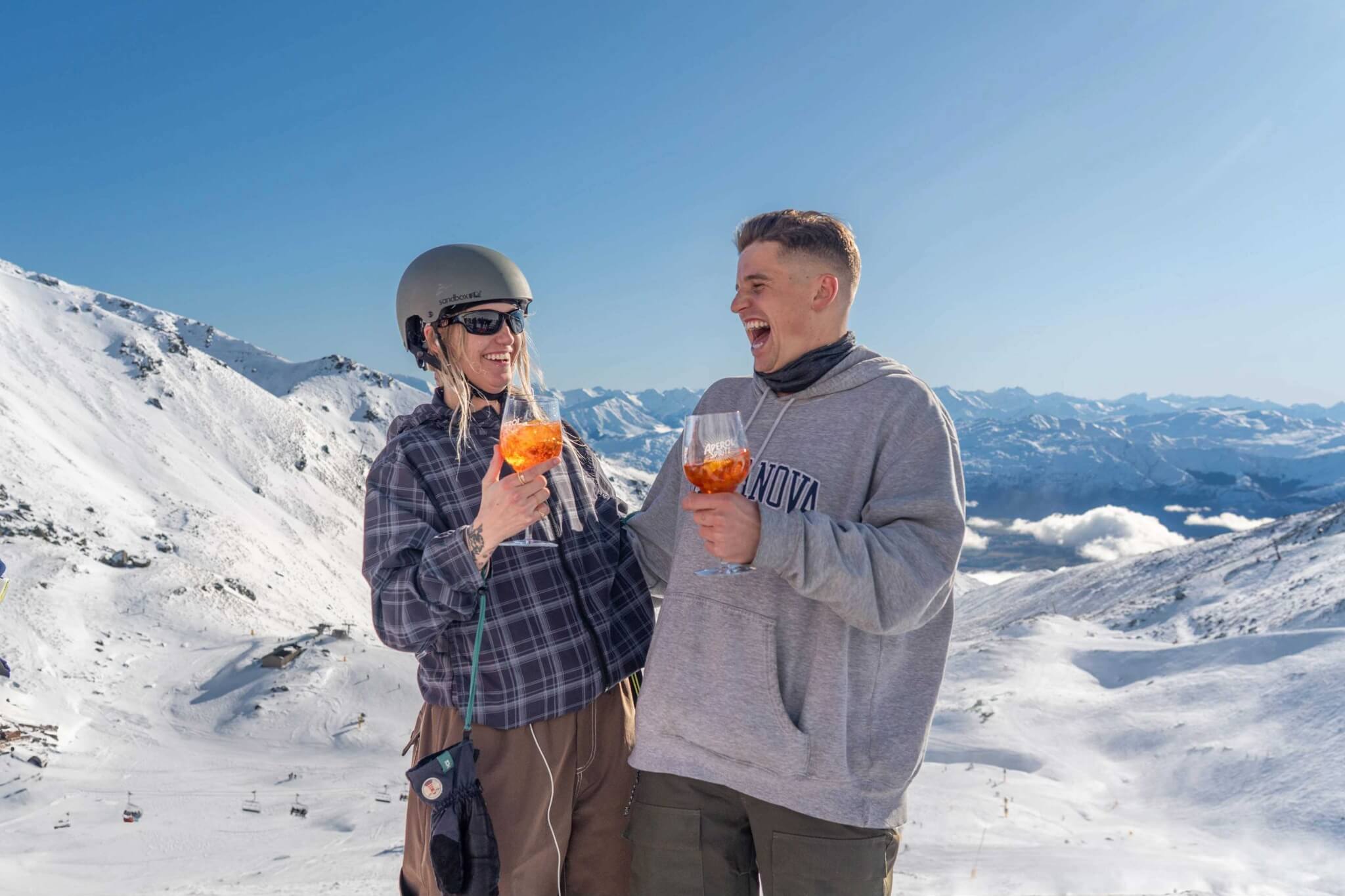 Two people in winter gear laughing and holding Aperol Spritz glasses with snowy mountain background.