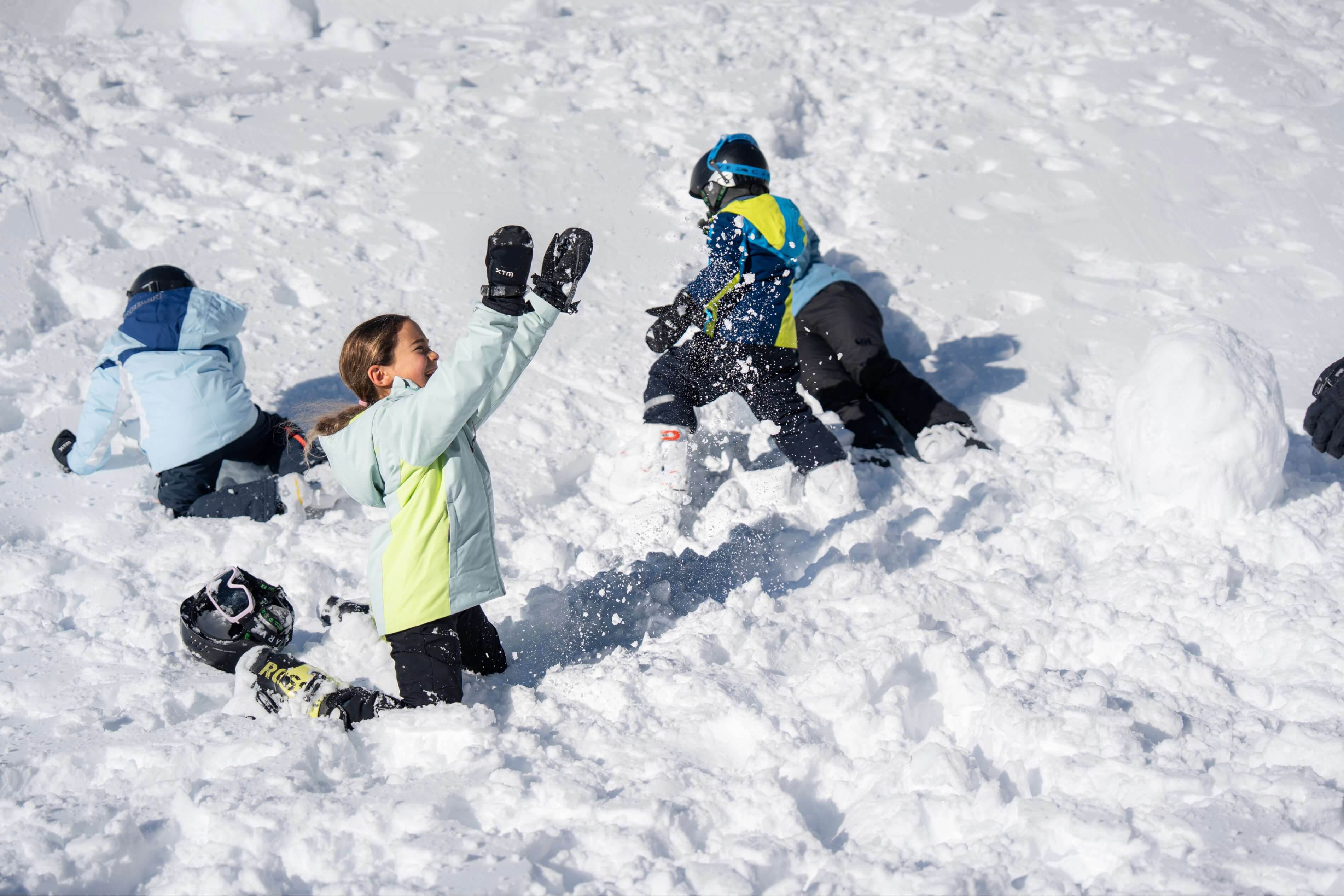 Children playing and throwing snow while kneeling in deep snow on a sunny winter day.