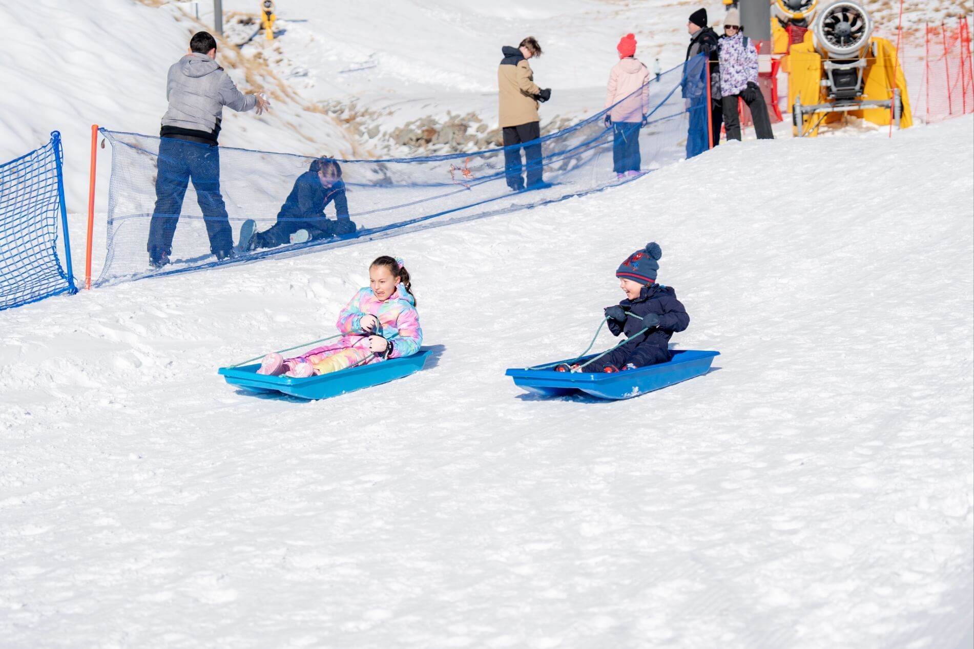 Two children sledding downhill on blue sleds in snowy area with people and netting in the background.
