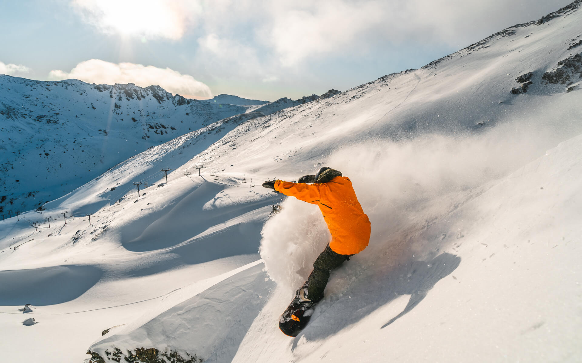 Snowboarder in orange jacket carving through fresh snow on a steep mountain slope with sunlight.
