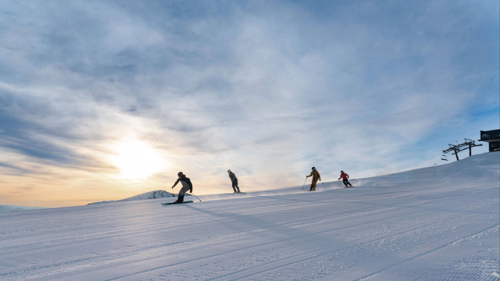 Four skiers descending a wide groomed snowy slope under a partly cloudy sky at sunrise.