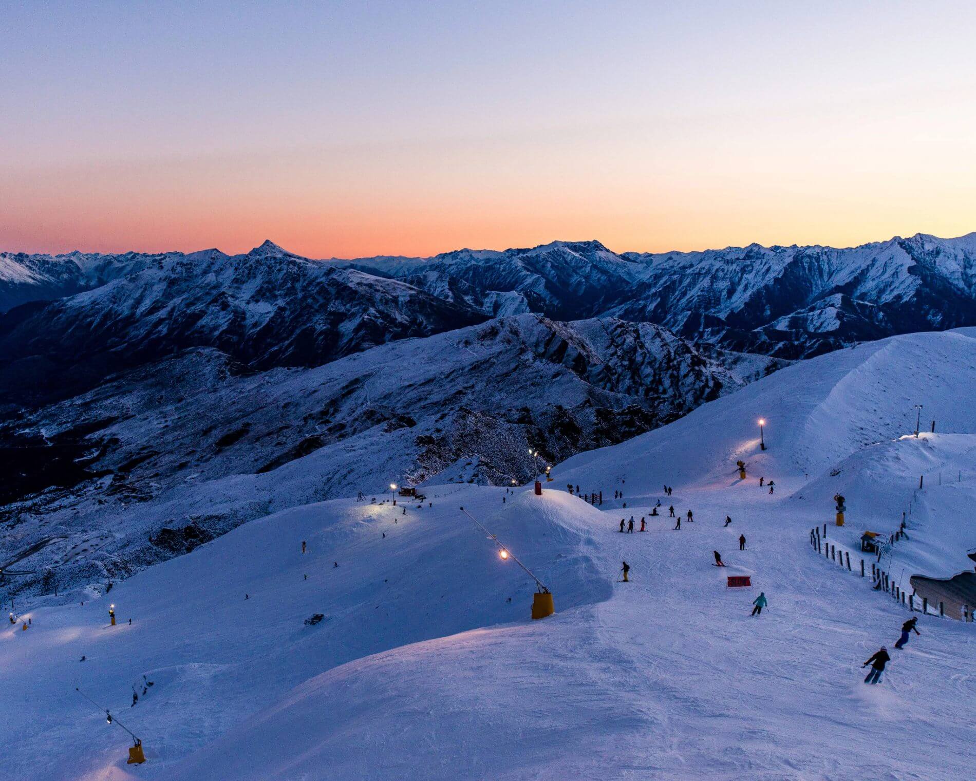 Skiers descend snowy slopes under lights at sunset with mountain range in the background.