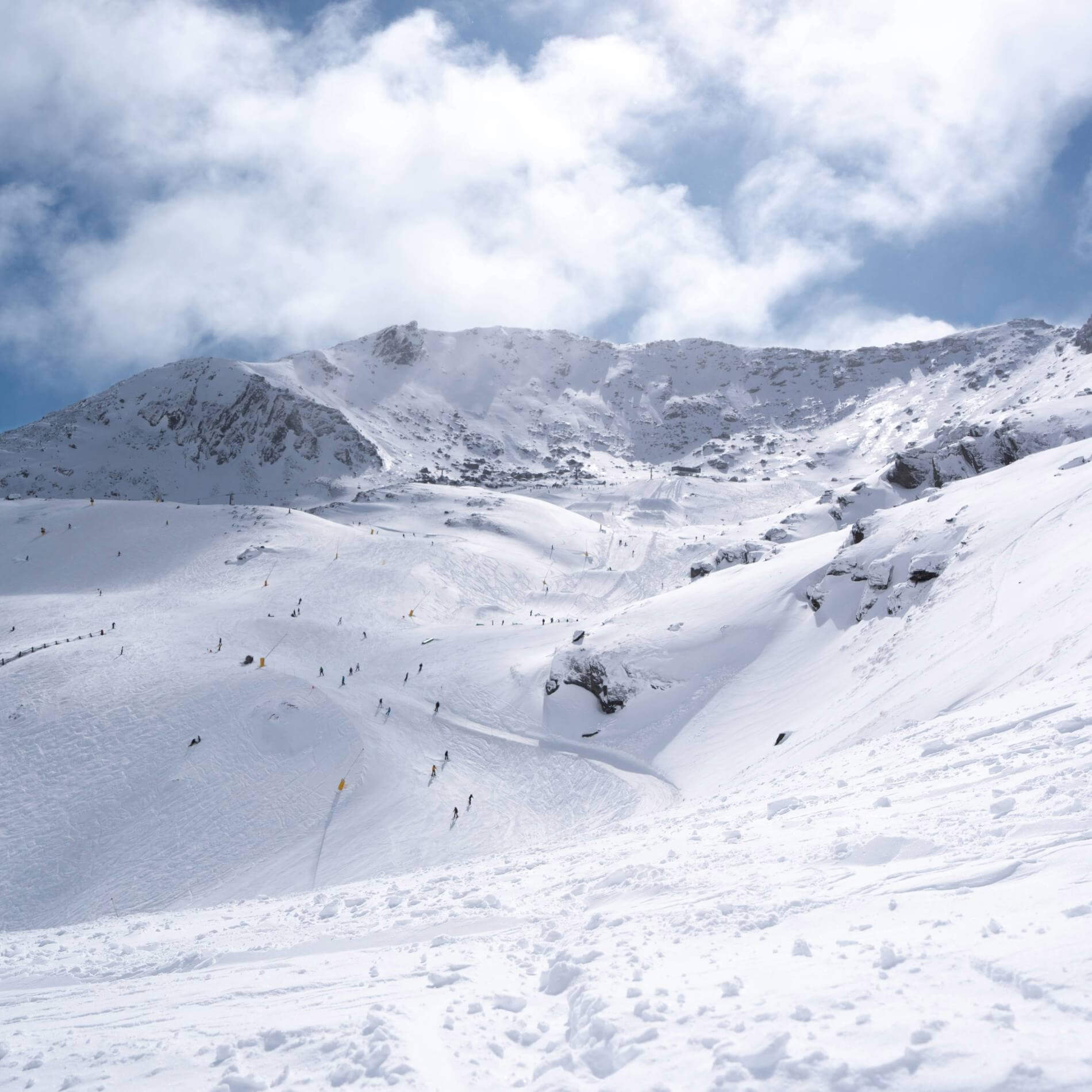 Wide snowy mountain slope with scattered skiers under a cloudy blue sky.